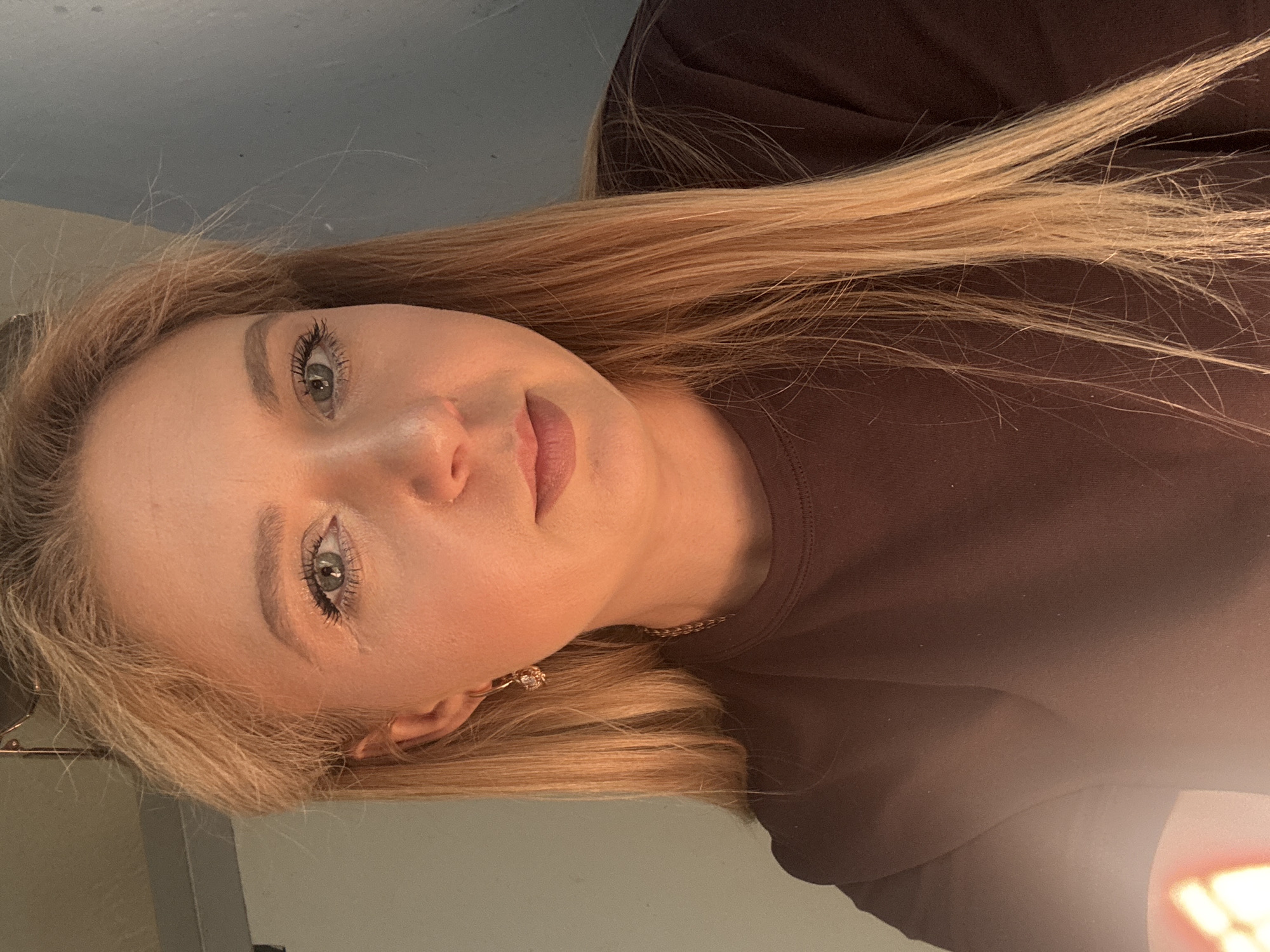 A blonde woman is shown from the chest up, facing the camera and looking slightly off to the left. She has fair skin and blue eyes. She is wearing a dark brown t-shirt and a gold chain necklace with a small charm. She has on a pair of small gold earrings with a clear gem. Her blonde hair is straight and falls past her shoulders. The lighting is warm, casting an orange hue on her face and chest. The background is dark and out of focus, with a hint of a blue wall on the right side. There is a bright light source in the lower left corner of the image, creating a lens flare effect.