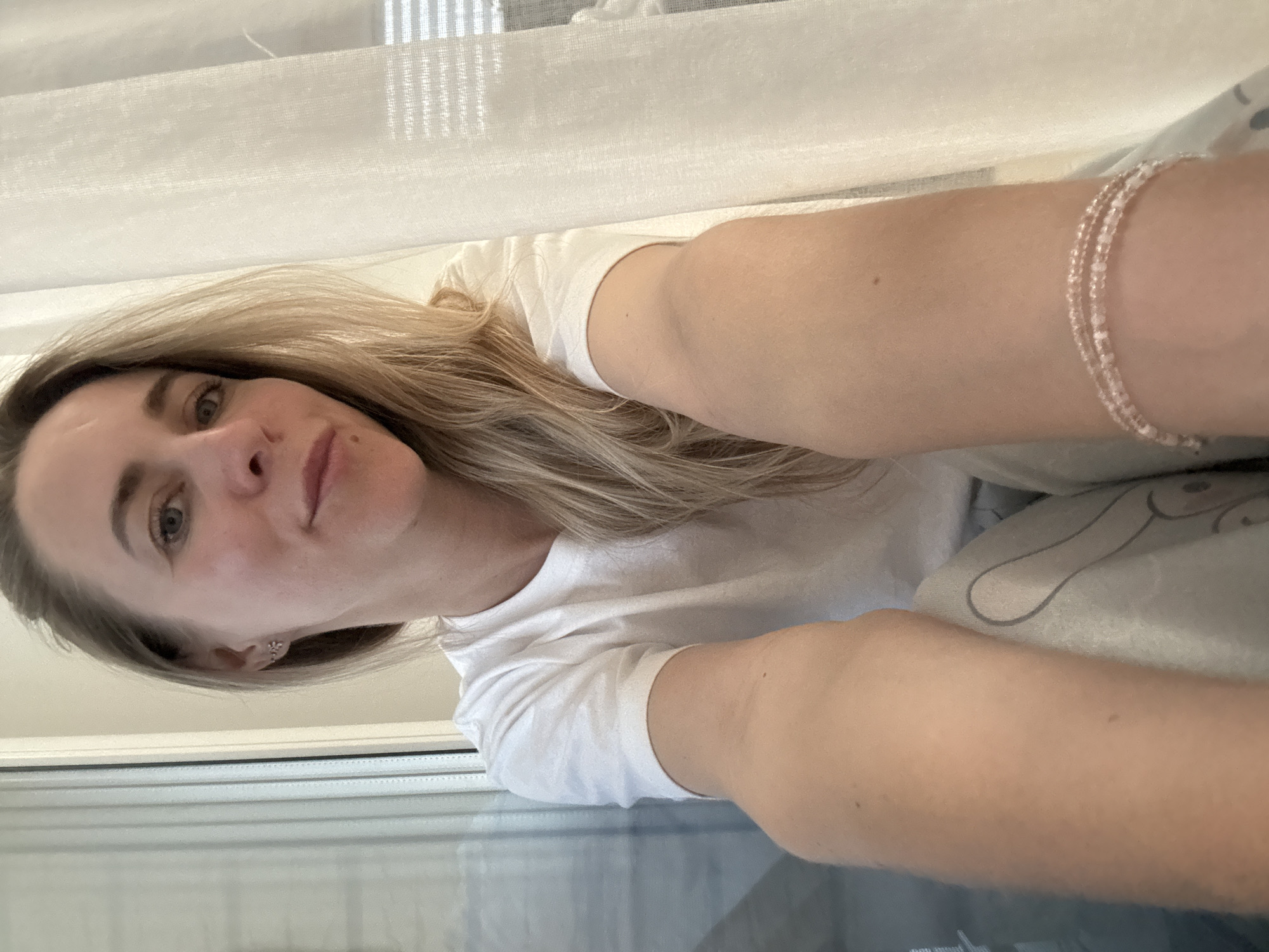 A selfie shot of a woman with blonde hair, wearing a white t-shirt and a bracelet, is oriented vertically. She appears indoors, with a window behind her. The specific location in the city of Chaikovsky, Russia, cannot be determined from the visual information provided.