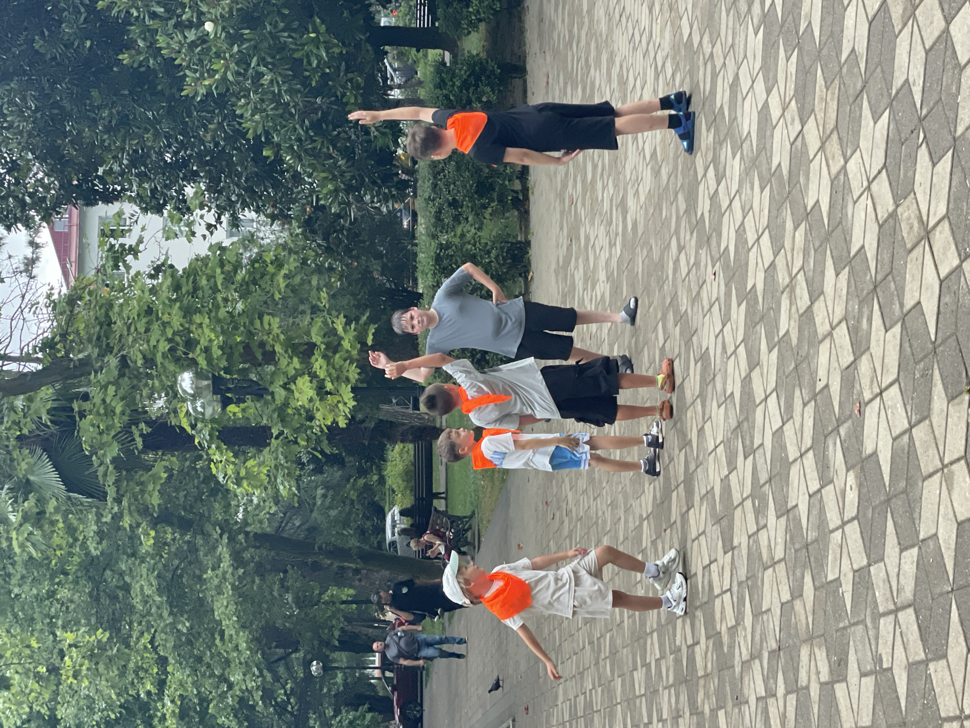 In an outdoor paved area, possibly a park or square in Sochi, Russia, five young boys are engaged in an activity. The boys are dressed in casual clothing, and four of them are wearing bright orange bandanas around their necks. One boy in the foreground, wearing a white t-shirt, light-colored shorts, and a white cap, is looking to his left with his arms extended. To his right, another boy in a white t-shirt and blue shorts, also wearing an orange bandana, has his arms raised. A third boy, in a grey t-shirt and black shorts, stands with his hands on his hips, looking towards the center of the group. A fourth boy, wearing a black t-shirt with an orange hood and black shorts, has one arm raised high. The fifth boy, in the background and to the left, is partially visible. In the background, there are trees, benches, and other people walking. The ground is paved with a patterned interlocking brick design. The overall atmosphere appears to be a recreational or playful gathering.