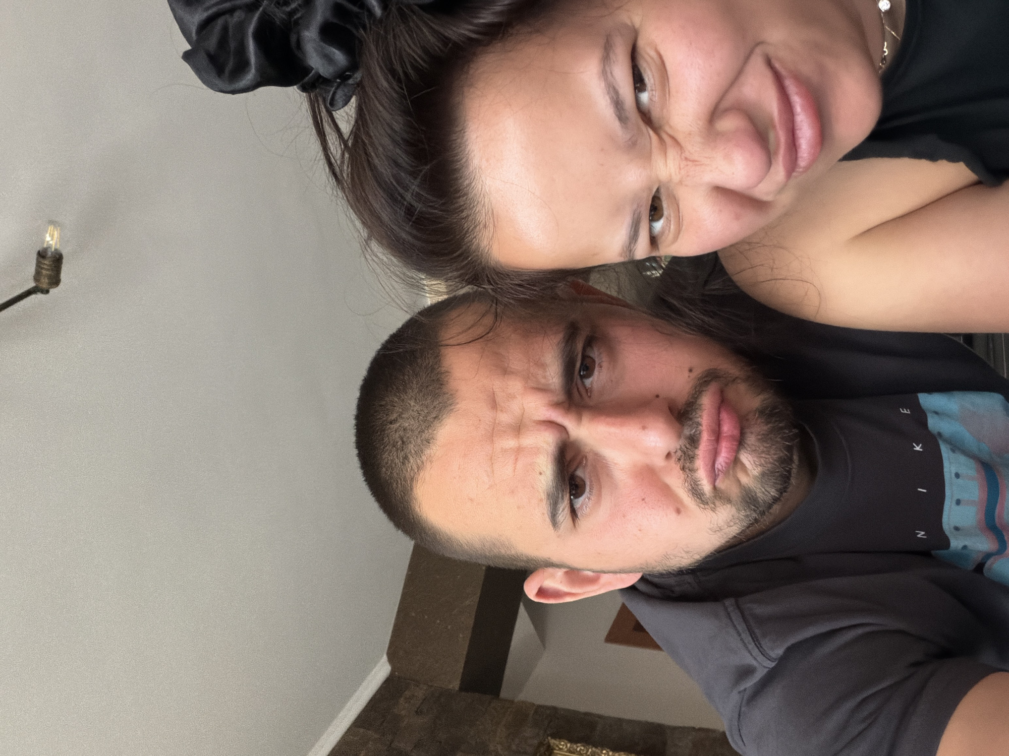 A selfie captures two individuals with expressive faces. The woman, positioned above, has dark hair with a black scrunchie and wears a black top. A gold necklace is visible around her neck. The man, positioned below, has short dark hair and a beard, wearing a black Nike t-shirt with blue accents and a gray jacket. They are both frowning. The background includes a neutral-colored wall with a lamp fixture. A partial view of a hallway with marble flooring is also visible. The location is Unknown, Unknown.