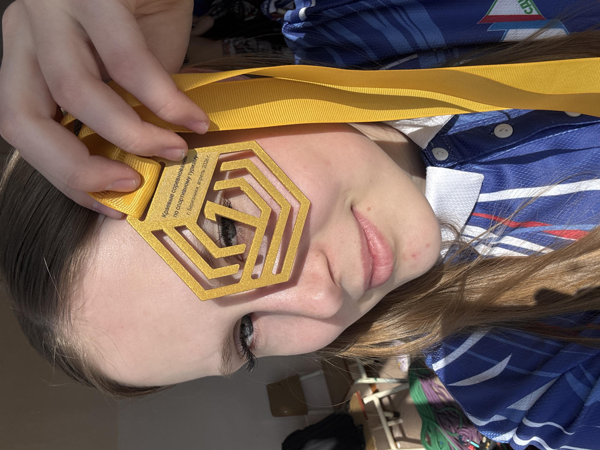 A close-up, angled shot depicts a woman holding a gold medal with a yellow ribbon in front of her face. The medal has a hexagonal design with geometric cutouts. Inscription on the medal reads, "Краевые соревнования по спортивному туризму, г. Березники, апрель 2026 г." indicating a regional sports tourism competition held in Berezniki, Russia, in April 2026. The woman has fair skin, light brown hair, and is wearing a blue and white sports jersey with buttons at the collar. A glimpse of a room interior is visible in the background.