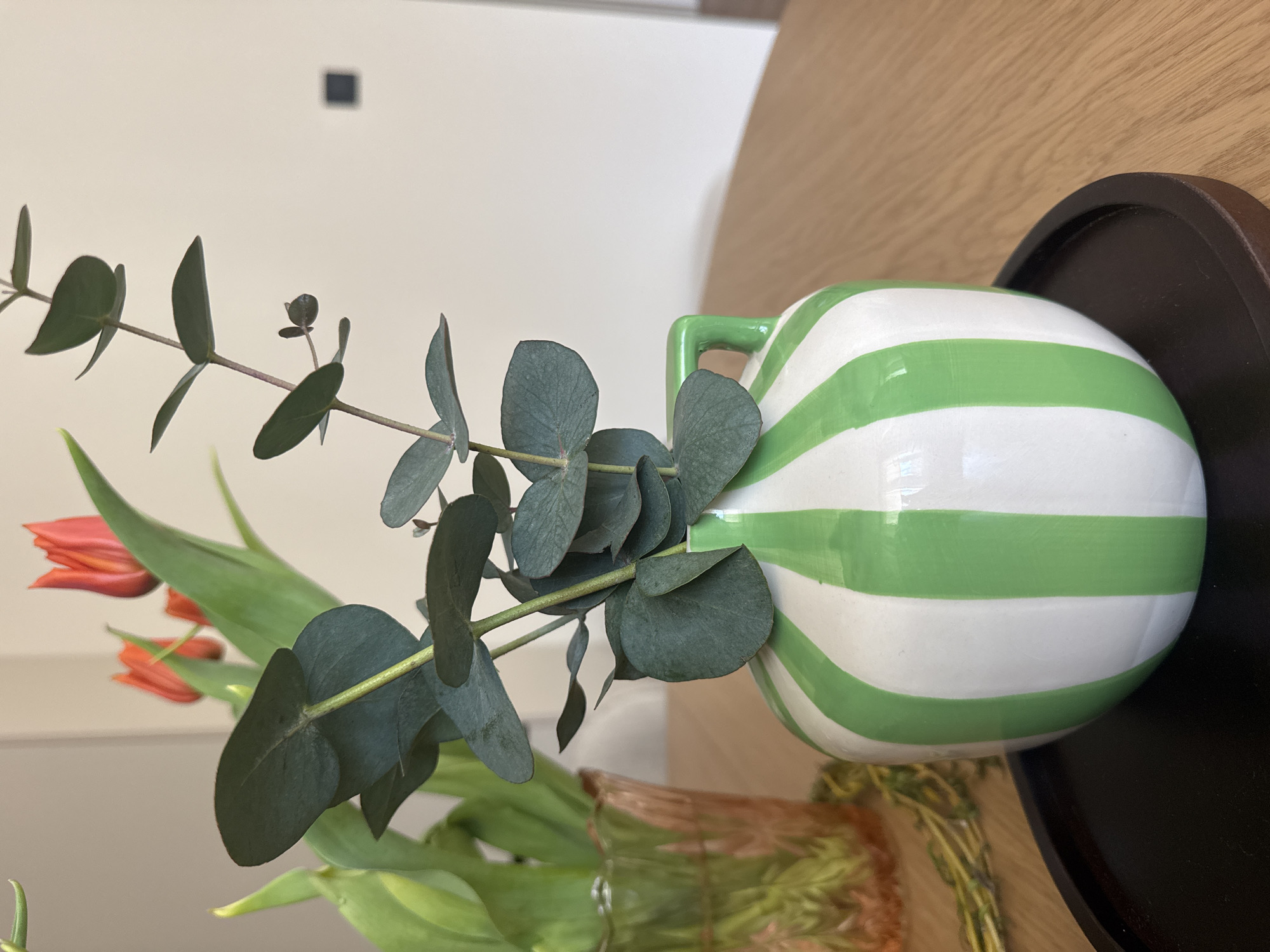 A close-up, eye-level shot shows a white vase with green horizontal stripes, sitting on a dark brown, shallow, round tray on a wooden surface. Inside the vase are orange tulips and eucalyptus branches. The vase has a green handle. The background includes a white wall with a small, dark square object and a blurred wooden surface. The scene appears to be indoors. There is no evidence to place this scene in Burgas, Bulgaria.