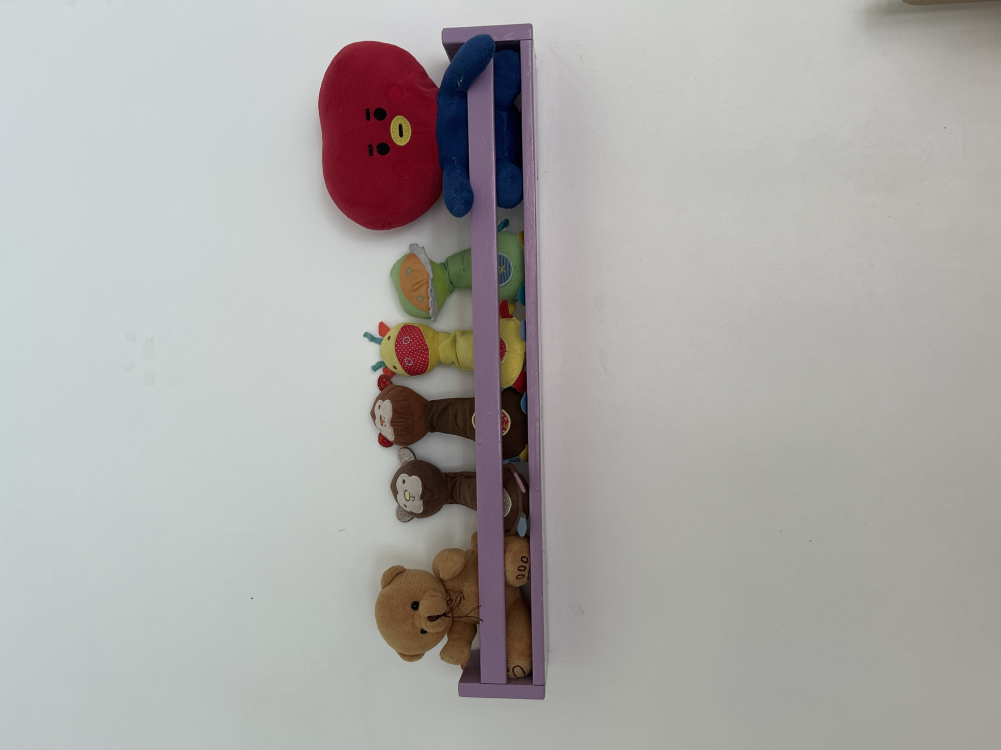 A purple shelf is mounted on a white wall. On the shelf are six stuffed toys. From left to right, the toys are a brown teddy bear, two monkey puppets, a yellow giraffe puppet, a green alien puppet, and a large red character puppet. The teddy bear is sitting and appears to be the largest of the toys. The monkey puppets are standing upright. The giraffe and alien puppets are also standing. The red character puppet is sitting and is the tallest. The background is a plain white wall.