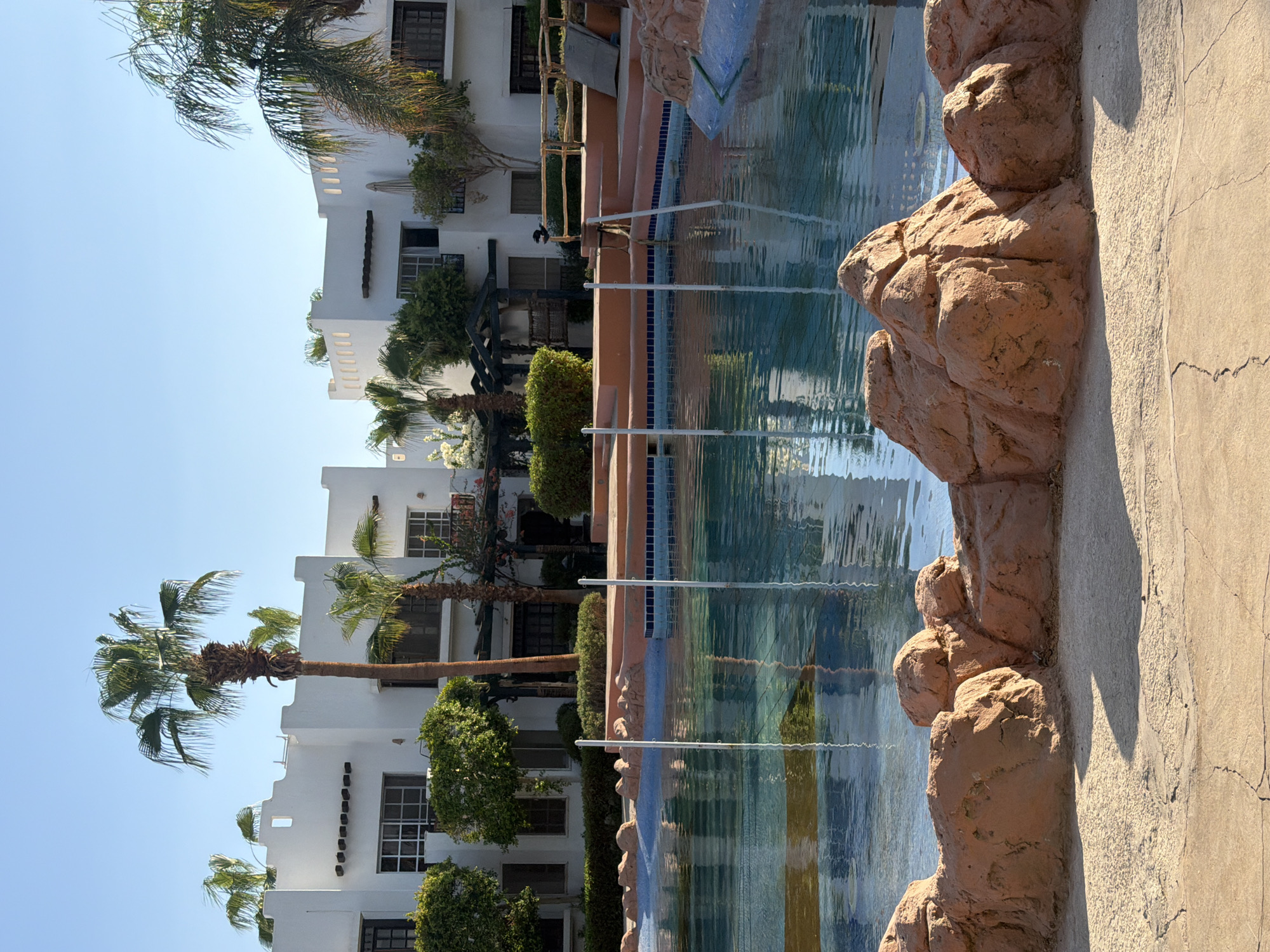 A vertical, eye-level shot shows a swimming pool and buildings in Sharm El Sheikh, Egypt. The clear blue sky is visible in the top half of the image, with the sunlit buildings and palm trees in the middle, and the shimmering turquoise swimming pool in the foreground. The buildings are white and have flat roofs and dark windows, with some palm trees and greenery surrounding them. The pool has a rocky, terracotta-colored edge and is filled with clear blue water that reflects the sky and buildings. Two white poles stand in the water, suggesting a swimming lane. The foreground is a sandy patio with cracks in it.