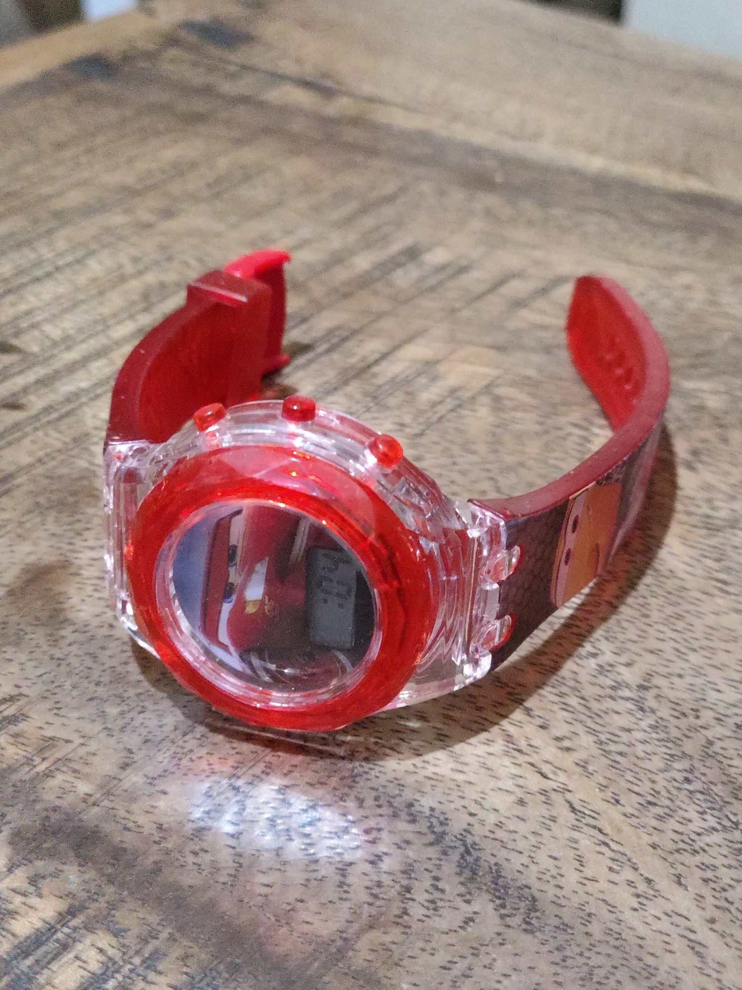 A red digital watch is lying on its side on a wooden surface. The watch has a transparent plastic case with red buttons on the top. The band of the watch is red and has cartoon characters printed on it. The watch face displays a digital time of 12:07. The wooden surface appears to be a table with a grainy texture. The lighting suggests an indoor environment. The specific location of Bulakrejo, Indonesia cannot be determined from the provided image.