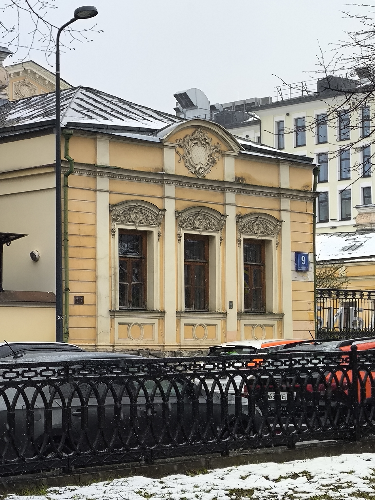 A daytime street scene in Moscow features a light yellow, two-story building with a classical architectural facade. This building is adorned with three ornately framed windows and a decorative cartouche above the central windows. Its mansard roof is partially covered with snow, indicating cold weather. A dark green downspout runs down the left side of the building, and a black lamppost stands prominently in the left foreground, with bare tree branches visible against the overcast sky.
A blue street sign with the number "9" and smaller Cyrillic text (likely "KAP") is affixed to the building's right side. In the background, a taller, modern-style building with numerous windows and rooftop ventilation units is visible. Another smaller yellow building is partially seen behind a fence on the right.
The foreground is dominated by a decorative black metal fence featuring intertwining oval and rectangular patterns. Behind this fence, several cars are partially visible, including a dark SUV or hatchback, an orange car, and a white car. The ground in front of the fence and along the street is dusted with snow.
Visible text and numbers include "38" on the lamppost, "9" and "KAP" on the street sign, and on a car's license plate, parts like "69 E" and "749 433". The overall atmosphere is quiet and cold under an overcast sky.