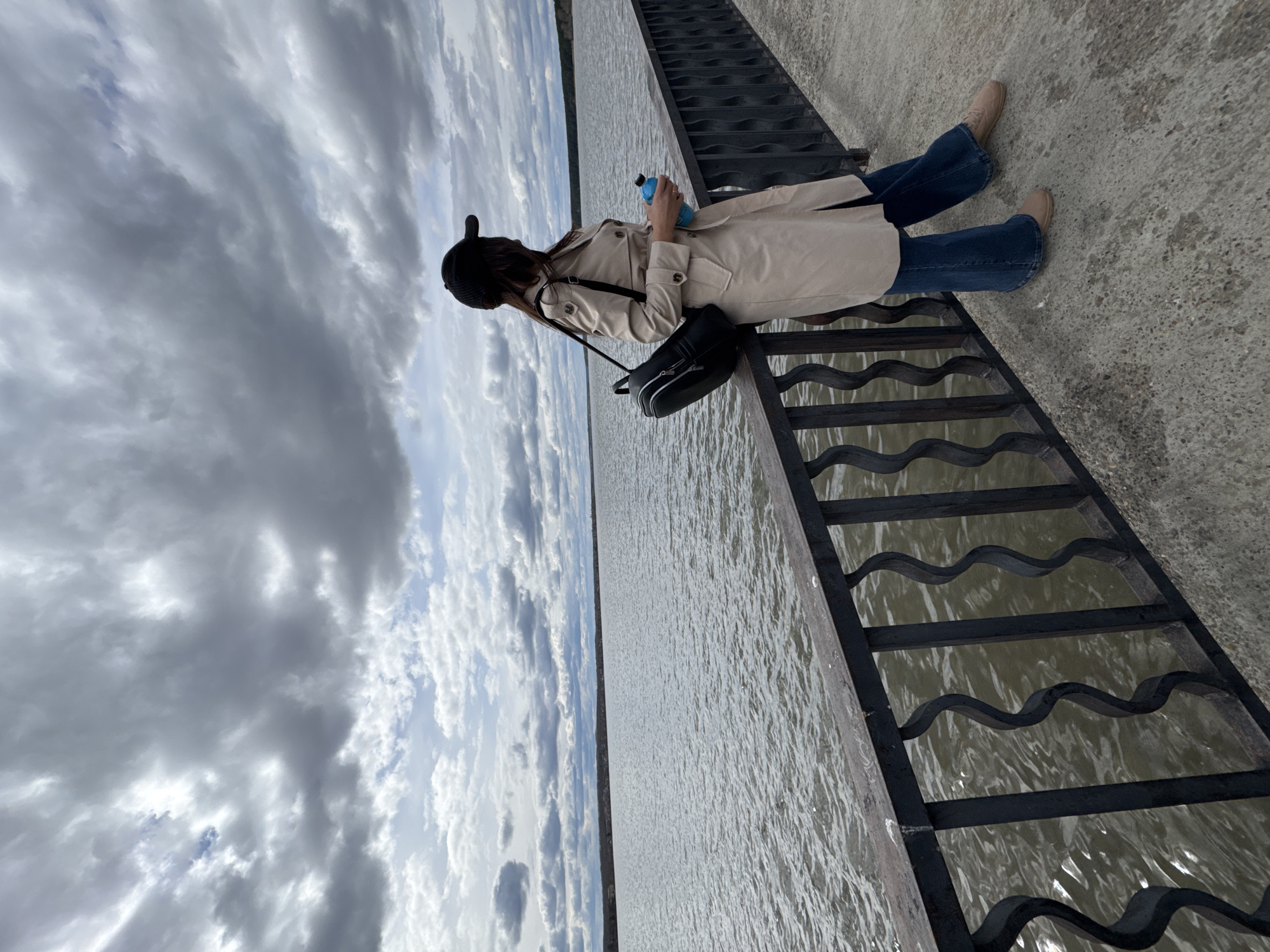 A woman sits on a concrete pier in Votkinsk, Russia. She wears a black cap, tan trench coat, blue jeans, and tan shoes. She holds a blue water bottle. A black purse hangs from her shoulder and rests on a black metal railing. The railing separates the pier from a body of water that stretches to the horizon under a cloudy sky.