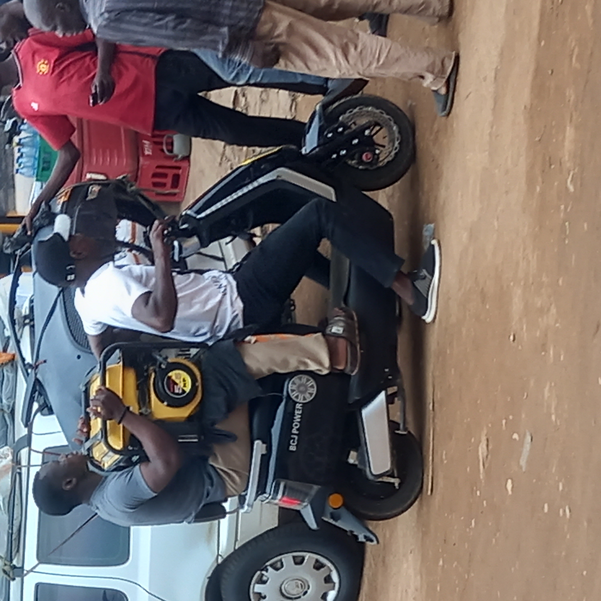 In Sardauna, Nigeria, the scene shows two men sitting on a black three-wheeled vehicle, labeled "BCJ Power," on a dirt ground adjacent to a white van. One man wears a gray t-shirt and is seated behind, the other wears a white t-shirt and a black cap. Additional people and a red crate are visible in the background.
