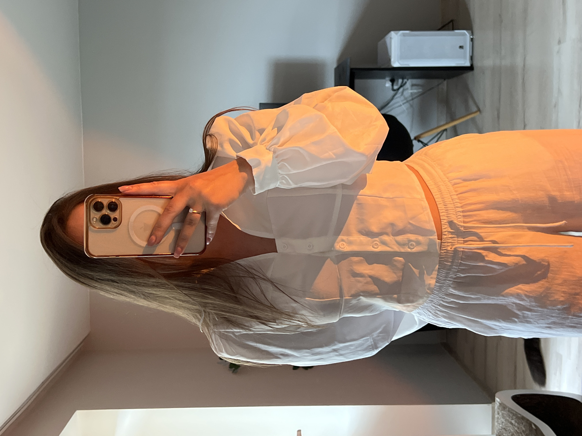 A vertical mirror selfie captures a person, likely female, from the chest to the mid-thigh, showcasing an all-white outfit in an indoor setting. The person holds a light-colored smartphone with a visible MagSafe ring and a metallic frame (possibly gold or rose gold) in their right hand, obscuring their face as they take the picture. A simple ring with a clear stone is worn on the ring finger of their left hand. Their long, light brown hair falls over their left shoulder.
The attire consists of a white collared shirt with prominent, puffed sleeves gathered at the cuffs and a button-down front. The shirt appears cropped or tied at the waist, revealing a narrow strip of midriff. Below, the person wears white shorts or loose-fitting pants with an elasticated, ruched waistband and a visible drawstring, suggesting a comfortable, casual style. Both garments have a light, slightly crinkled texture.
The background features a light-colored wall, possibly off-white or light gray. To the left, a dark shelf or desk is visible, holding a white, rectangular object that could be a small appliance or speaker. Some wires and cables are discernible behind the shelf. The lower part of the background shows what appears to be wooden flooring or wall paneling. The overall lighting has a warm, yellowish cast, illuminating the white clothing. No discernible text is visible on any objects or clothing. The image does not contain any specific visual cues to indicate its location as Ternopil, Ukraine.