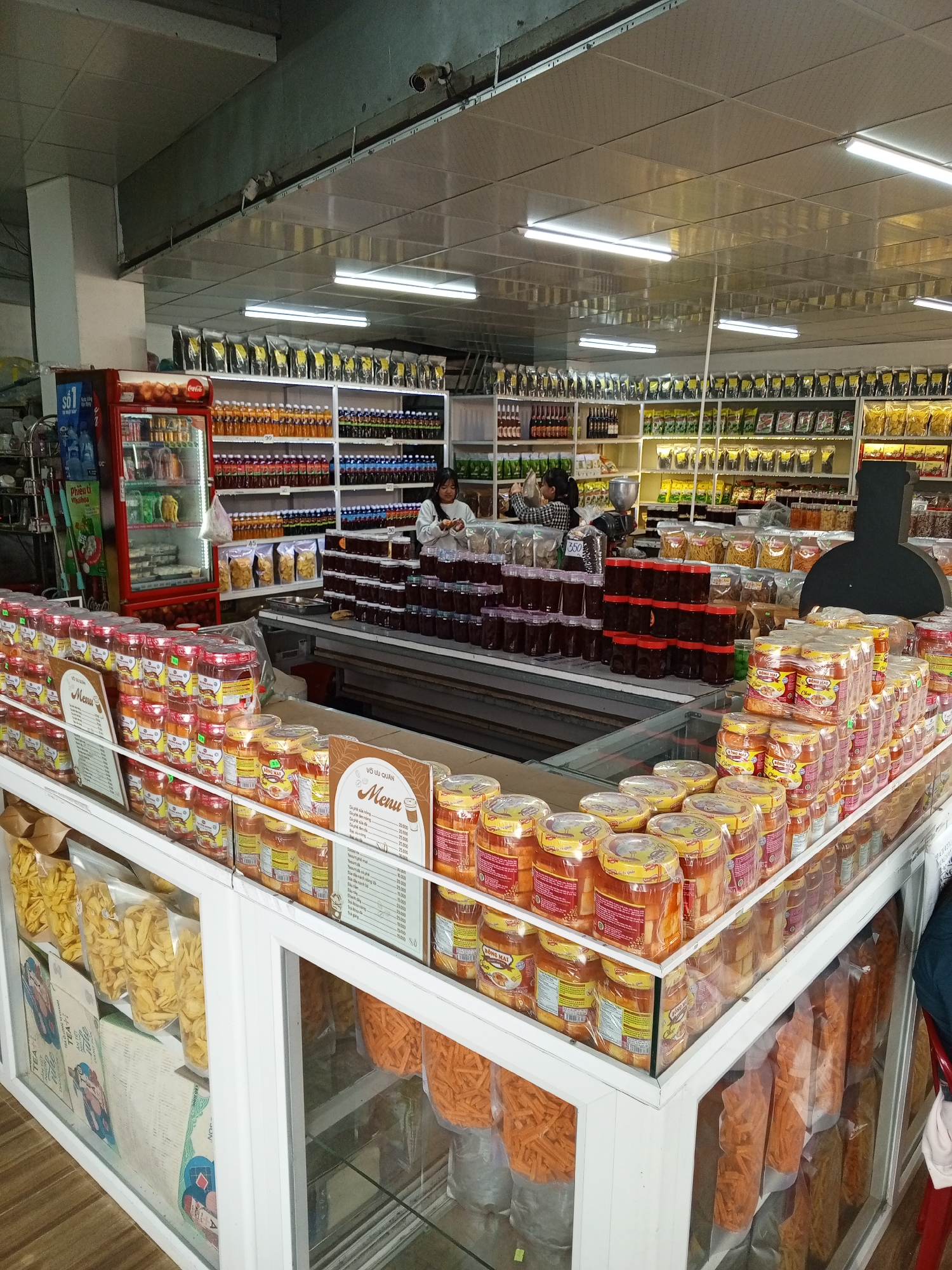 This is a photograph of a brightly lit convenience store or small market, likely in Xuân Thành, Vietnam, filled with rows of merchandise. The foreground features a display case filled with various packaged food items, including bags of yellow chips and large jars of preserved foods or sauces. The items are organized and neatly arranged. Behind the display case, more shelves are stacked with a diverse selection of goods. In the middle ground, there is a refrigerated display cabinet containing bottled drinks. Further back, the store extends with extensive shelving units stocked with numerous products, including bottled beverages, packaged snacks, and other food items. Two individuals are visible in the background; one is a woman standing at a counter and the other is partially visible. The overall environment is clean and organized, typical of a retail establishment. The lighting is bright and consistent throughout the space, likely from overhead fluorescent fixtures.