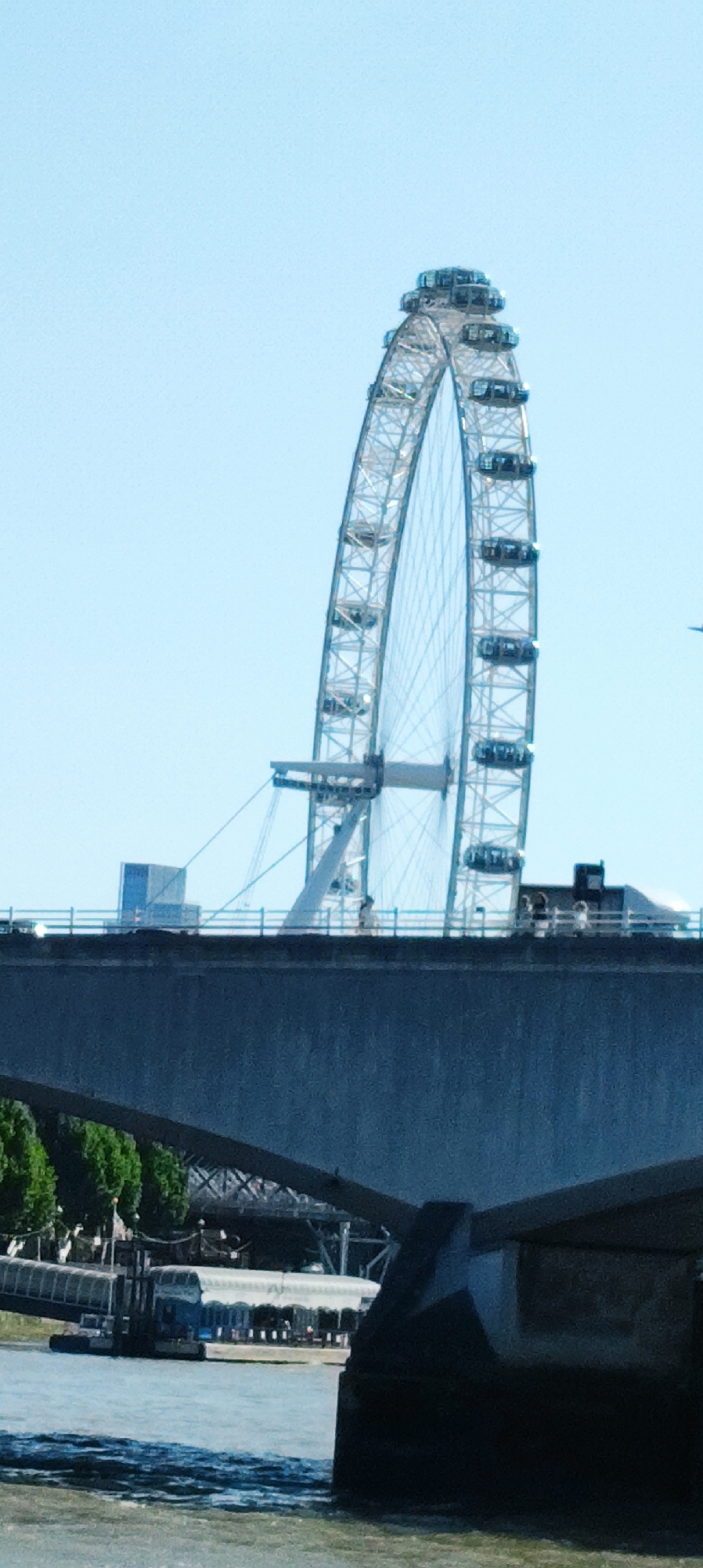 The image shows a view of the London Eye, a large Ferris wheel, in London, United Kingdom. The London Eye is a prominent landmark and tourist attraction. The image is taken from a low angle, looking up at the Ferris wheel. In the foreground, a bridge is visible, with parts of the river and a boat also in view. The sky is clear and blue, with no clouds. The London Eye is in operation, with several pods visible on the wheel. There are people on the bridge, and some appear to be on the London Eye itself. The overall scene is one of a bustling city day.
