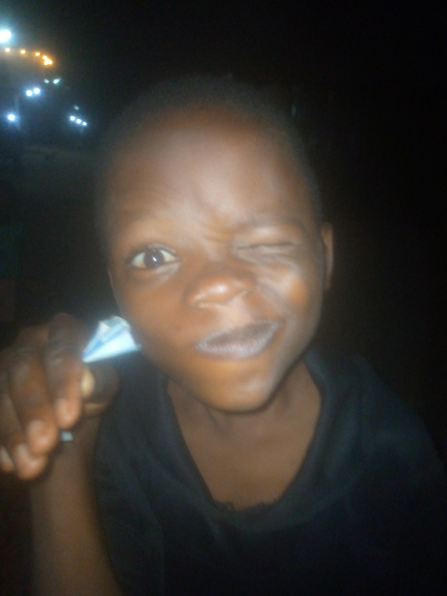 A close-up photo shows a person of African descent with dark skin, wearing a dark garment. They are squinting one eye. The background is dark, but lights are visible in the upper left. The subject is located in a city of Unknown, Unknown. The person is holding a small, folded white and blue object in their fingers, pointed towards the viewer.