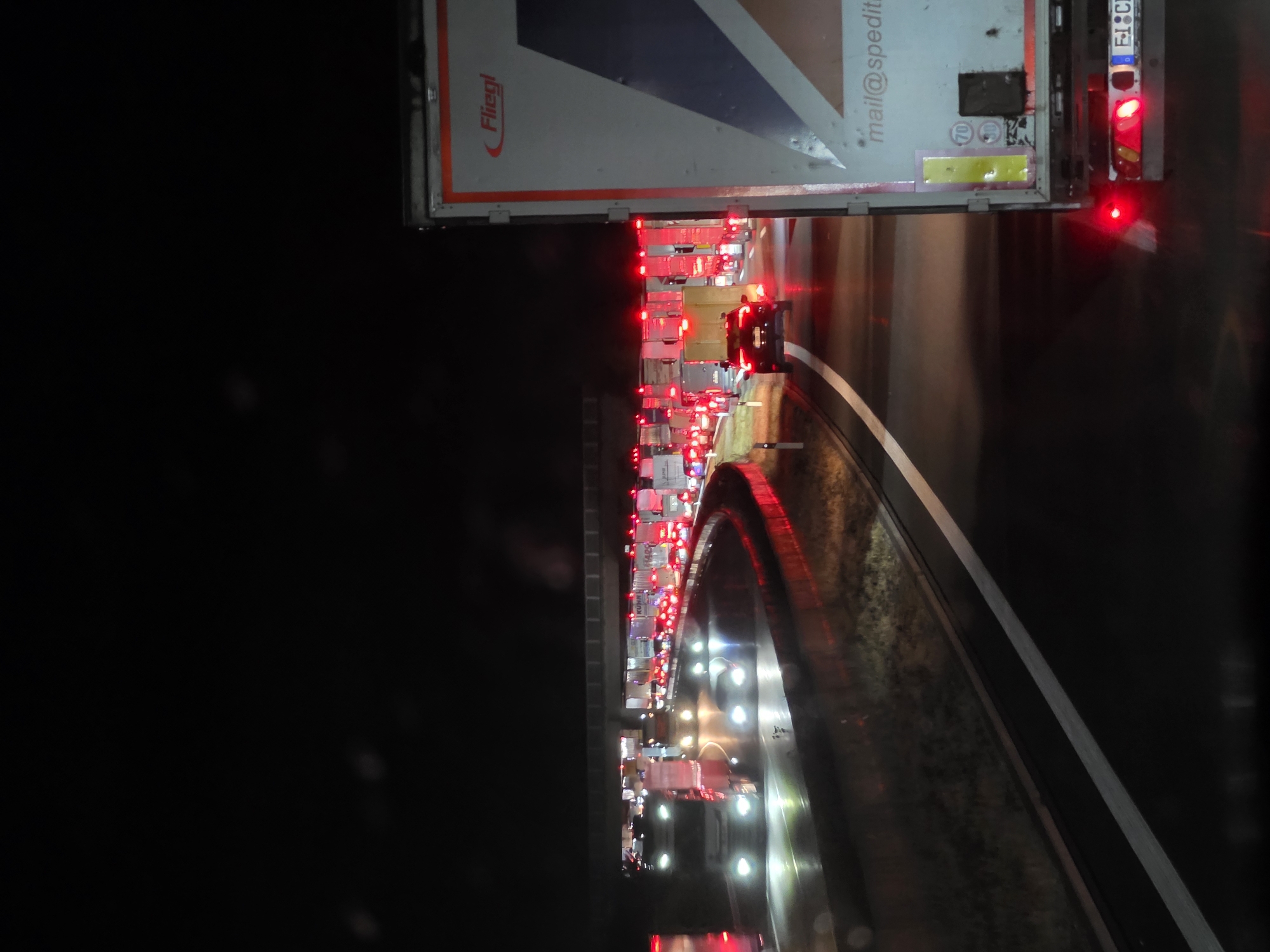 The image shows a traffic jam at night on a highway in Uettingen, Germany. Numerous trucks and cars are stopped, with their brake lights illuminated. The road appears wet, suggesting recent rain. In the foreground, a large truck with "Fliegl" branding is visible, and its rear lights are on. The sky is dark, and the only light sources are the vehicle lights and overhead streetlights. The scene conveys a sense of standstill and congestion.