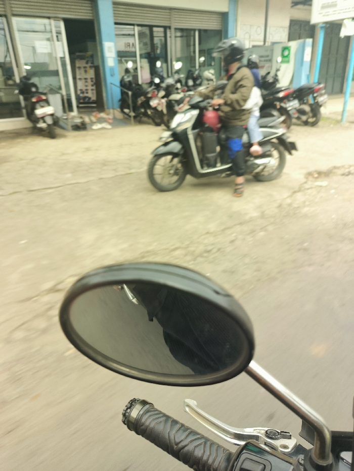 The image shows a rear view mirror of a motorcycle traveling on a road in Ciparay, Indonesia, located at latitude: -7.029, longitude: 107.697. The motorcycle's mirror reflects another motorcycle in the distance, driven by a man wearing a helmet and a dark jacket. There is a second person on the back of the motorcycle, obscured by the reflection. The image does not show any other people or objects. The road is asphalt and appears to be in good condition. The environment appears to be urban, with buildings visible in the background.