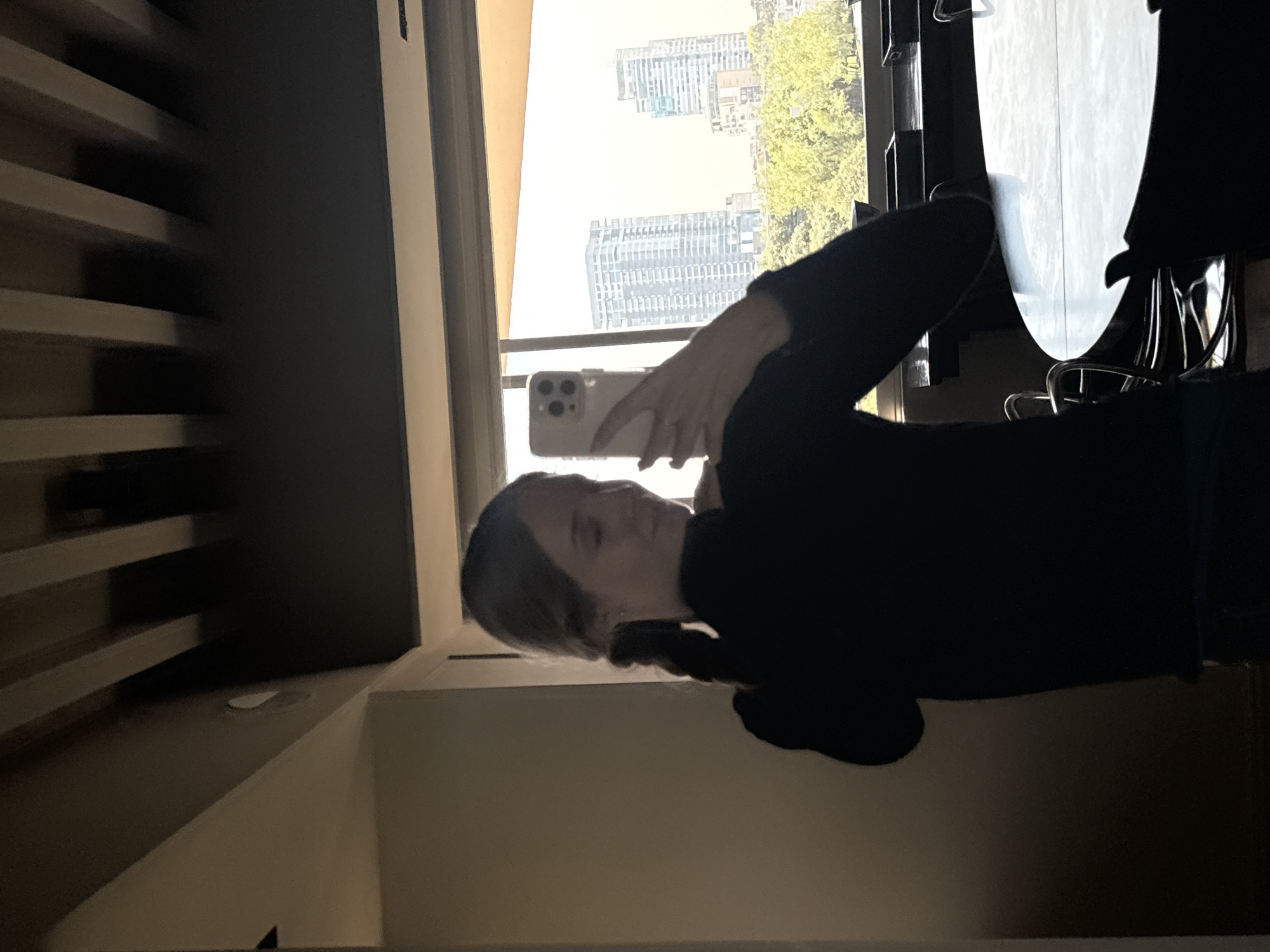 The image, originally rotated 90 degrees counter-clockwise, depicts an indoor scene with a person taking a selfie. A woman with light brown hair, wearing a dark long-sleeved top, is positioned in the foreground, largely silhouetted against a brightly lit window. She holds a white smartphone, likely an iPhone with multiple rear cameras, in her right hand, angled towards her face as if capturing a photo. Her face is partially obscured by the phone and shadows, showing a neutral expression.
To the right of the woman, a segment of a round, light-colored table with dark chairs is visible. On the left, dark, slatted furniture, possibly shelving, stands against a wall.
Through the expansive window or reflective surface, a clear view of a modern city skyline is apparent. Several tall, contemporary buildings and skyscrapers are visible under a bright, sunny sky. Lush green trees are also prominent in the outdoor scene, indicating urban greenery. The stark contrast between the bright outdoor light and the dimmer indoor setting creates a strong backlighting effect on the interior subjects. No visible text is present in the image. The setting is a modern urban environment, aligning with the provided location context of Mexico City, Mexico.