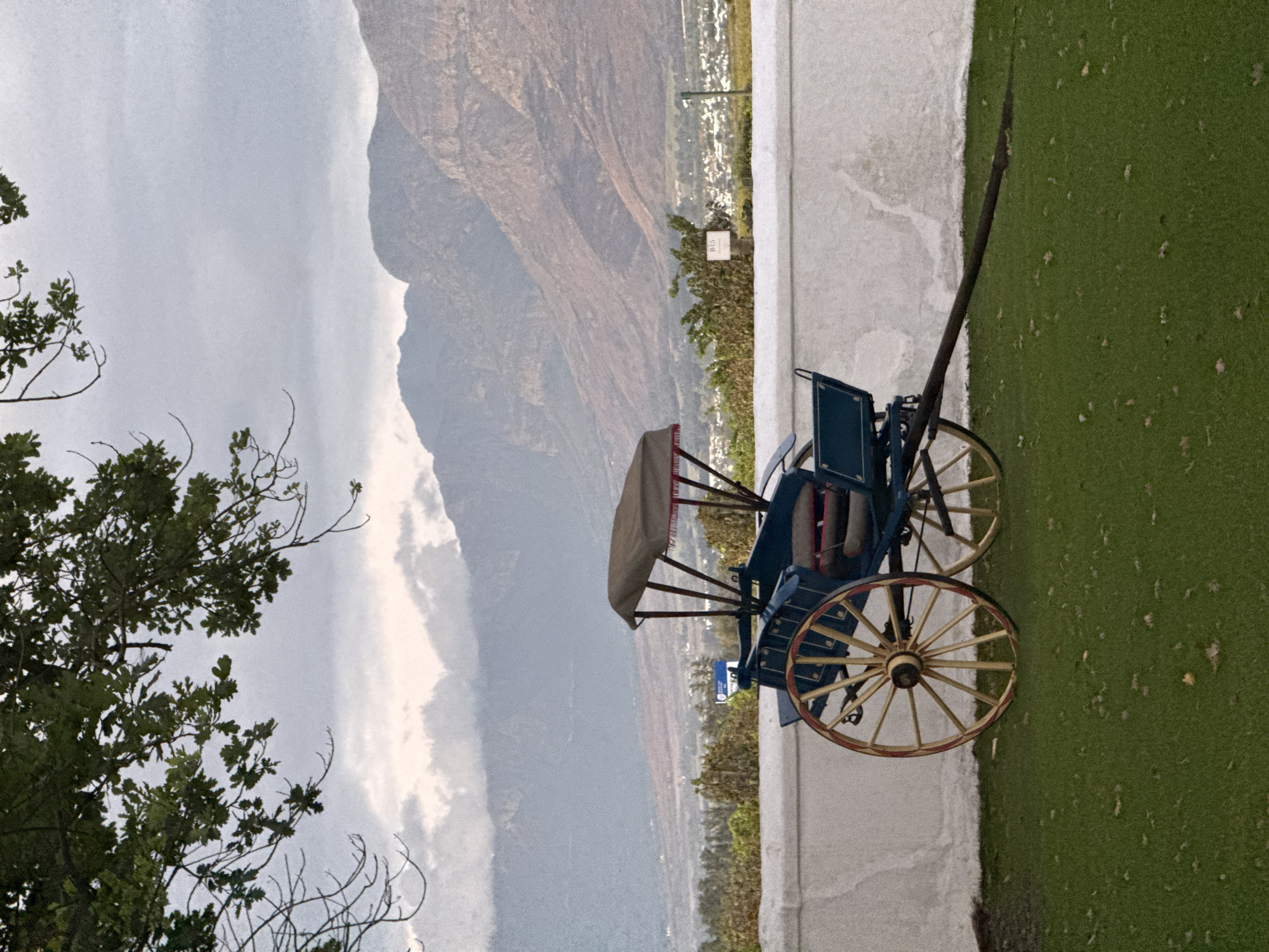 The image displays a panoramic landscape featuring a vintage carriage in the foreground against a backdrop of majestic mountains and a valley. The sky is overcast or hazy, covering the top portion of the frame. Below, rugged mountains with shades of brown and grey stretch across the mid-ground, appearing vast and imposing. A valley, possibly home to a small town, is discernible at the foot of the mountains.
In the immediate foreground, a well-manicured lawn of green grass extends from the right, meeting a low, white wall or barrier. A striking blue, two-wheeled carriage with intricate red detailing is positioned on the grass. Its large, spoked wheels are painted yellow with red rims, and it features a light-colored canopy over the seating area. A long, dark shaft protrudes from the front of the carriage.
To the left, a large tree with dense green foliage frames the scene, partially obscuring the sky and distant mountains. A small white sign bearing the text "B15" is visible in the mid-ground, tucked amidst smaller greenery behind the white wall, below the mountains.
No specific activity or event is taking place; the carriage is stationary, suggesting it may be a decorative element or a prop. The diffused lighting indicates an overcast day or perhaps late afternoon. The overall impression is one of tranquility and natural grandeur, consistent with the Drakenstein Nu region of South Africa, known for its scenic mountainous landscapes.