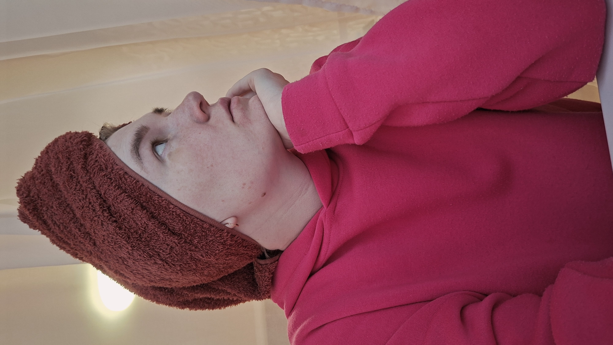 The image shows a person with light skin wearing a maroon towel on their head and a bright pink hoodie. The person is facing upwards and slightly to the left, with their hand touching their chin. Their face has freckles and possibly a mole on the neck. The background is a pale, possibly white, color, with a light source visible in the bottom left. There is no information to determine that this image was taken in Chaikovsky, Russia. The location cannot be definitively identified from the image.