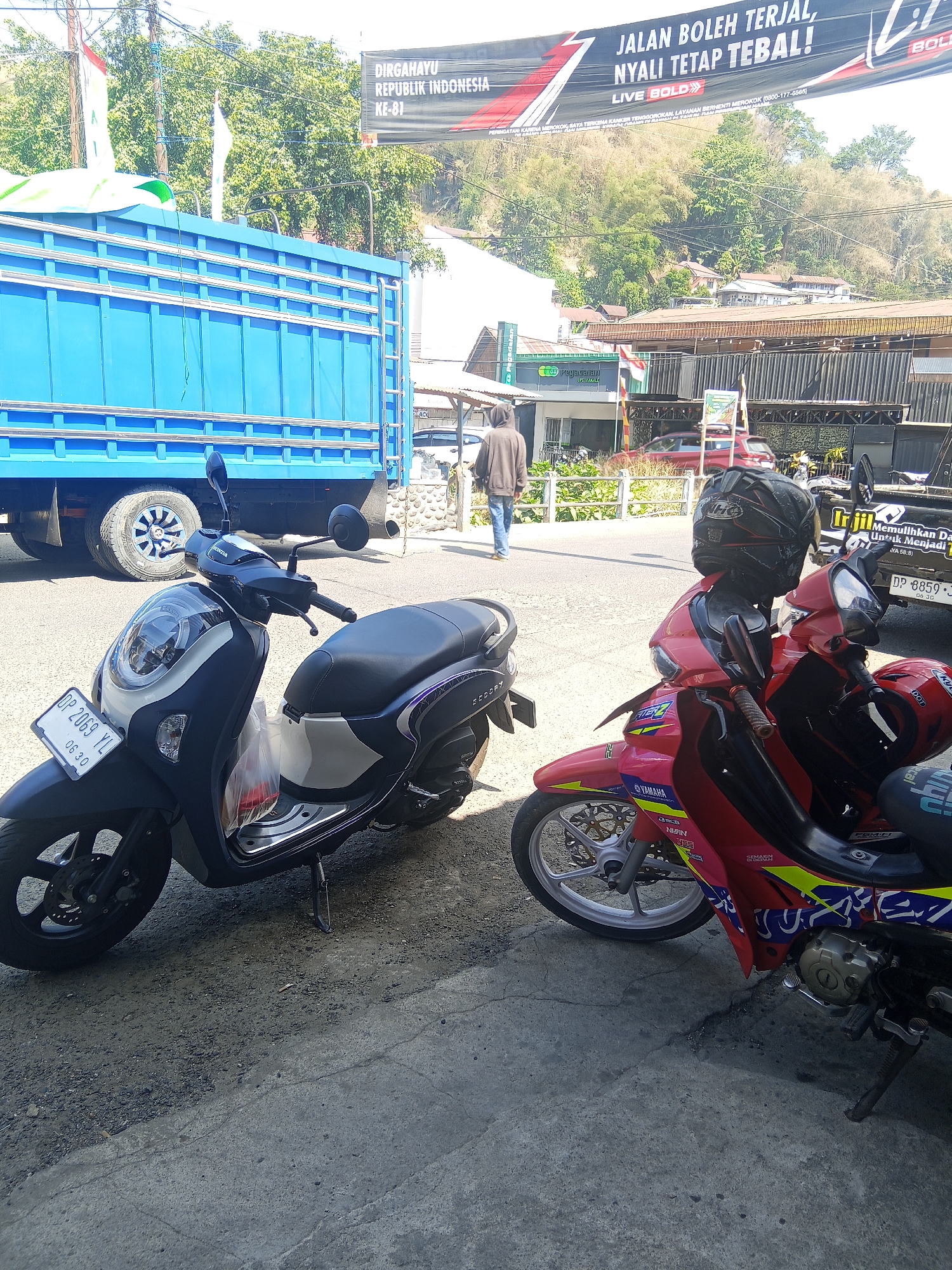In Makale, Indonesia, a street scene unfolds under a bright sky, featuring two parked motorcycles in the foreground. A black and white Honda scooter, bearing the license plate "DP 2069 YL" and the date "06 30", is positioned to the left. To its right, a vibrant red motorcycle is parked, with a black helmet resting on its handlebars. Behind the motorcycles, a large blue truck is visible, partially obscuring a row of buildings. In the background, a banner with Indonesian text, possibly related to a national celebration, hangs across the street. A person wearing a brown hoodie and jeans walks away from the camera on the sidewalk. The environment appears to be a commercial or busy area, with trees and hills forming the backdrop.