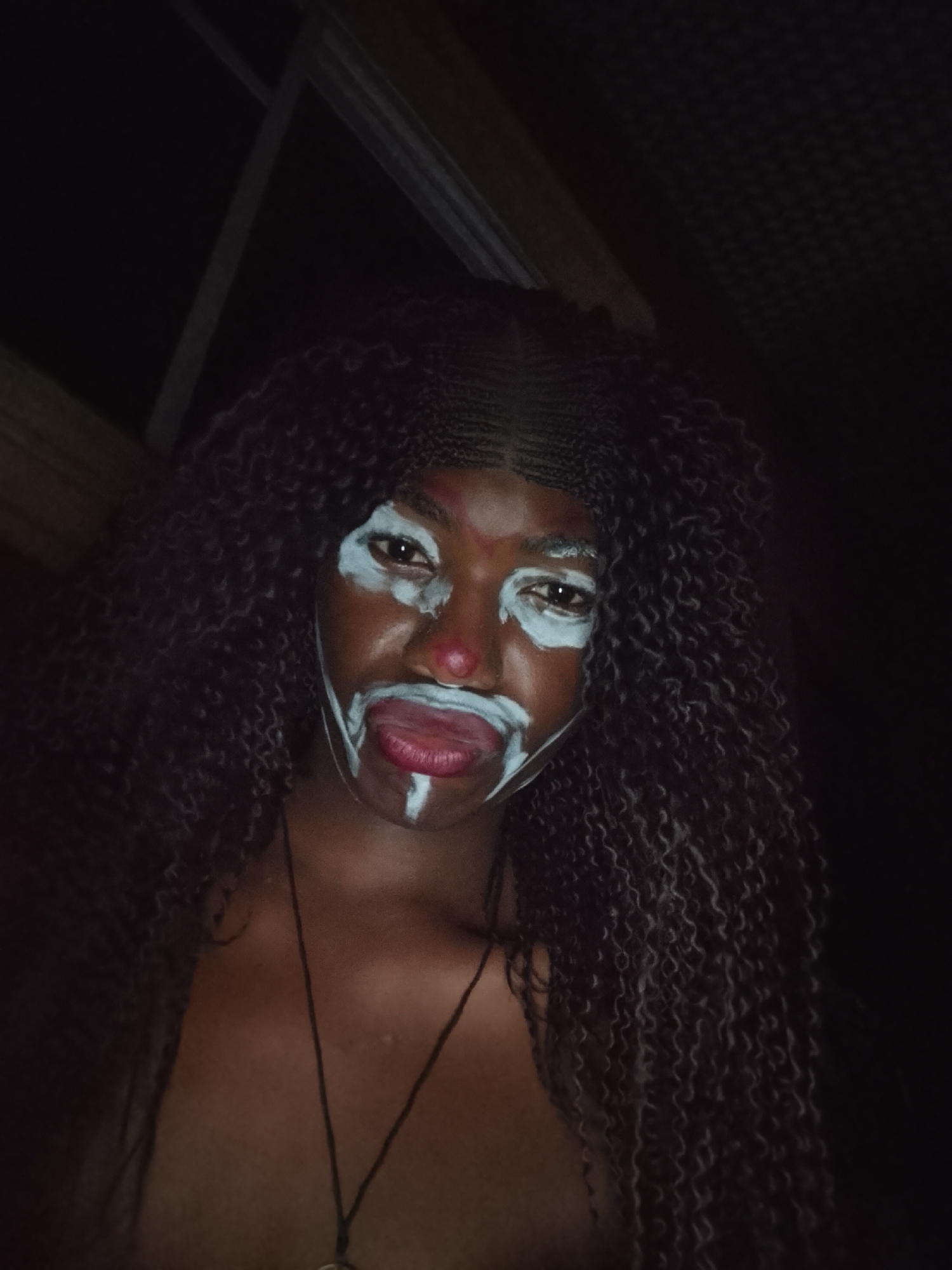 A low-angle, close-up shot shows a person with dark skin and long, dark curly hair, posing with their tongue out. They are wearing white face paint around their eyes and mouth, with a red dot on their nose. A black cord necklace is visible on their chest. The background is dark, with a glimpse of what seems to be a ceiling or roof structure in the upper portion of the frame. The location is unknown.