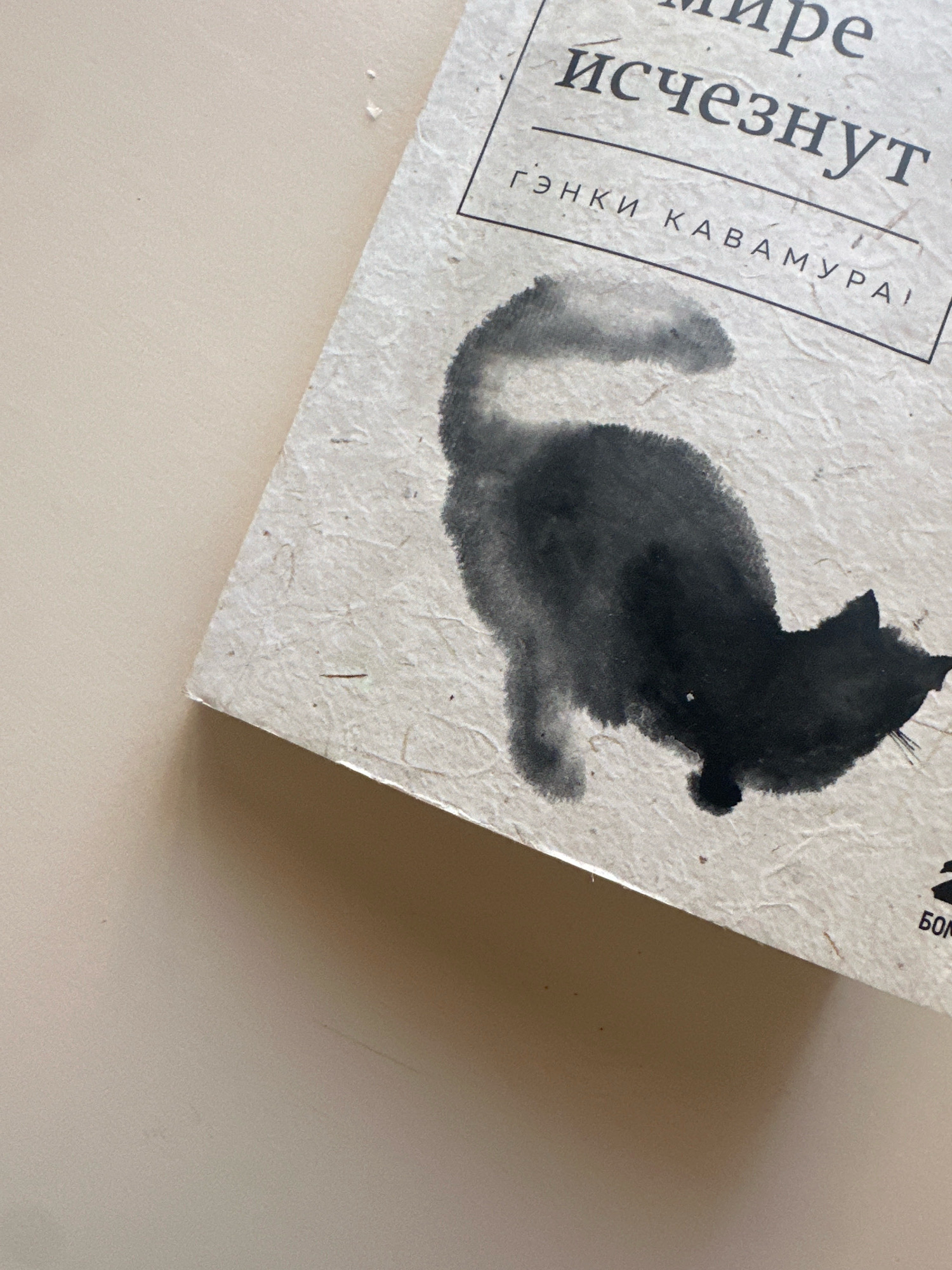 A close-up, overhead shot shows a portion of a book cover lying on a light beige, smooth surface, possibly a table. The book cover is made of a textured, off-white paper with visible fibers and small flecks. The cover features a black ink painting of a cat in a traditional East Asian style, with its back to the viewer and its tail curved upwards. The cat appears to be walking or looking to the right. Above the cat painting, black text in Cyrillic script is visible, part of a title and author's name. The text reads "и исчезнут" and below it "ГЭНКИ КАВАМУРА". The bottom right corner of the book cover shows a small portion of a black logo or symbol with the Cyrillic letters "БОМ". The lighting suggests a soft, natural light source from the top left, casting a subtle shadow on the beige surface to the right and bottom of the book. There are no people or any indication of the city of Gyumri, Armenia, in this image.