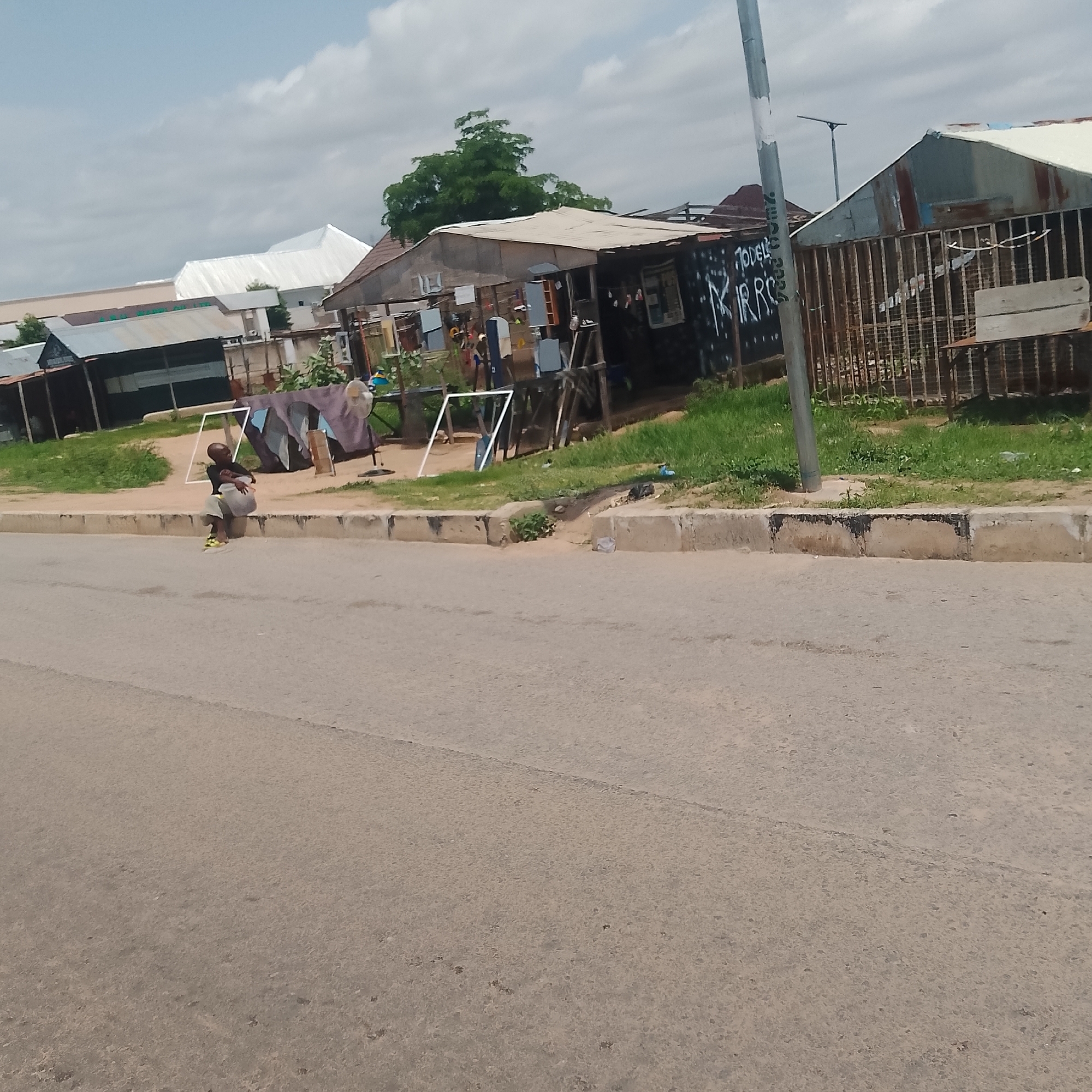 A young boy sits on a concrete curb in front of a row of market stalls in Jalingo, Nigeria. He is wearing a dark t-shirt, light-colored shorts, and yellow and green sneakers. The boy is curled up, with his arms wrapped around his legs. He is looking down. Behind him, the market stalls are constructed from corrugated metal and wood. One stall has graffiti on its side that reads "TODELI RR RO." Clothes hang on a rack in front of another stall. A large white fan is visible to the right of the stalls. The ground is covered in grass and dirt, with a small drainage channel running through it. A paved road runs in front of the curb. The sky is cloudy.