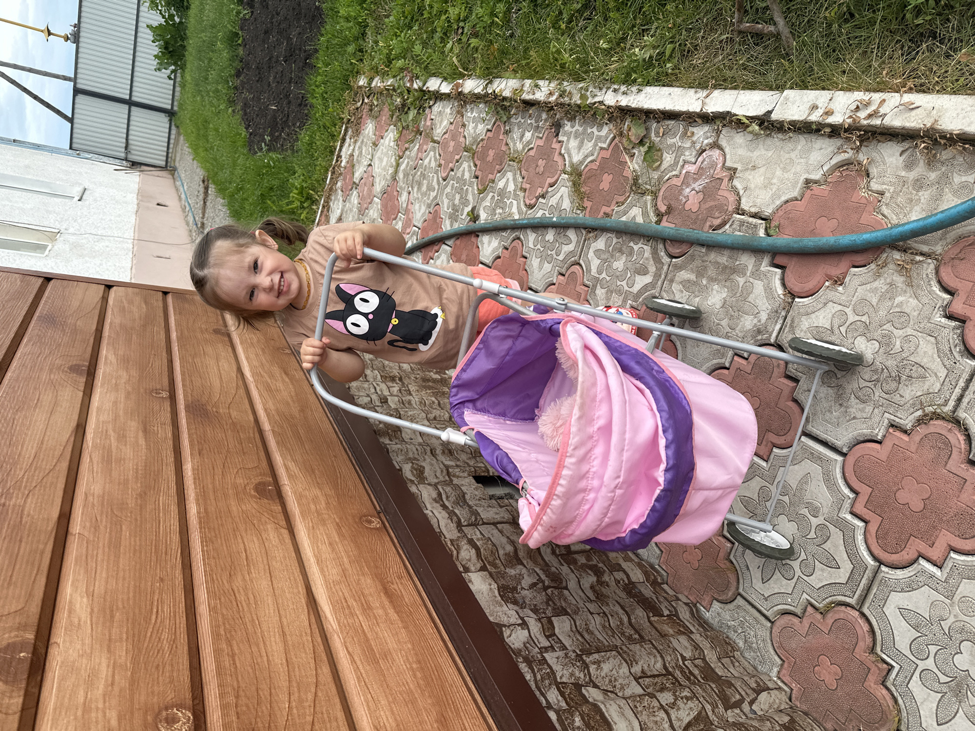 A young Caucasian girl, with blonde hair in two pigtails, wears a tan t-shirt with a black cat graphic and orange shorts. She stands outside a wooden-sided building, pushing a pink and purple doll stroller. The stroller has a metal frame and two wheels visible. The ground is paved with decorative interlocking tiles, and to the right, a green garden hose lies on the grass next to a patch of dirt. A white building with a light pink trim is visible in the background, along with a corrugated metal fence. The overall setting appears to be a residential yard in the village of Yaroslavka, Russia.