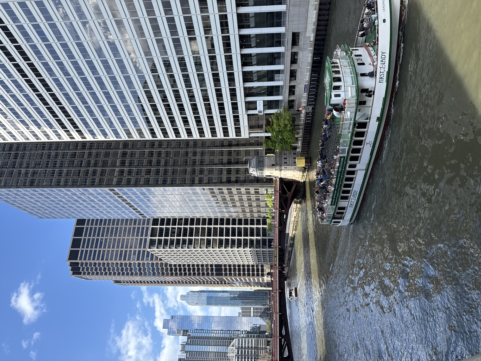 The image captures a view of the Chicago River in downtown Chicago, Illinois, USA. The "First Lady" river tour boat is seen navigating the waterway, carrying passengers. The architectural landscape is dominated by tall office buildings with grid-like window patterns. A bridge, possibly the Adams Street Bridge, crosses the river. The sky is partly cloudy.