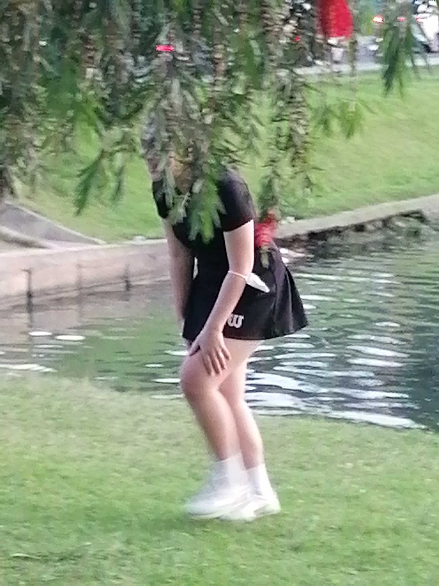 A person wearing a black skirt and white shoes is standing on a grassy area next to a body of water. The person's hands are placed on their thighs. The person is partially obscured by green foliage. The scene is outdoors.