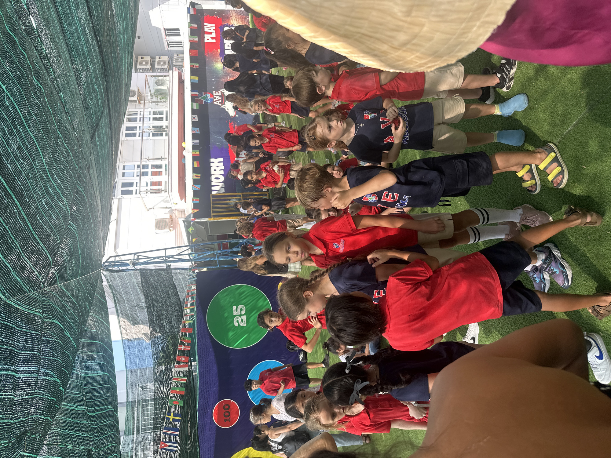 The image captures an outdoor event, possibly a school fair or sports day, on a sunny day. A green artificial turf surface covers the ground. In the background, a large banner displays the words "WORK", "AVE", and "PLAY" in large, colorful letters, with a graphic of a white dog or wolf. Smaller flags of various nations are strung across the top of the banner. A metal structure, likely part of a tent or stage, is visible on the right side.
The foreground and midground are filled with children and some adults, many of whom are wearing red or dark blue shirts and dark shorts or skirts, suggesting school uniforms. Some children have their backs to the camera, while others are facing forward. One child in the mid-left is wearing a red shirt and dark shorts. Another child in the foreground on the left is partially obscured, showing a tanned shoulder and arm.
Several circular targets with numbers are visible on a dark blue backdrop behind the crowd. One red circle contains the number "100", and a large green circle contains the number "25". A few individuals are standing near these targets.
The overall atmosphere appears to be one of a lively gathering, with children milling around and engaging in activities. The presence of the banner with "WORK", "AVE", and "PLAY" suggests an educational or recreational purpose for the event. The lighting and shadows indicate it is daytime. The specific location in the city of Nam Nha Trang Ward, Vietnam, is not visually evident from the image itself.