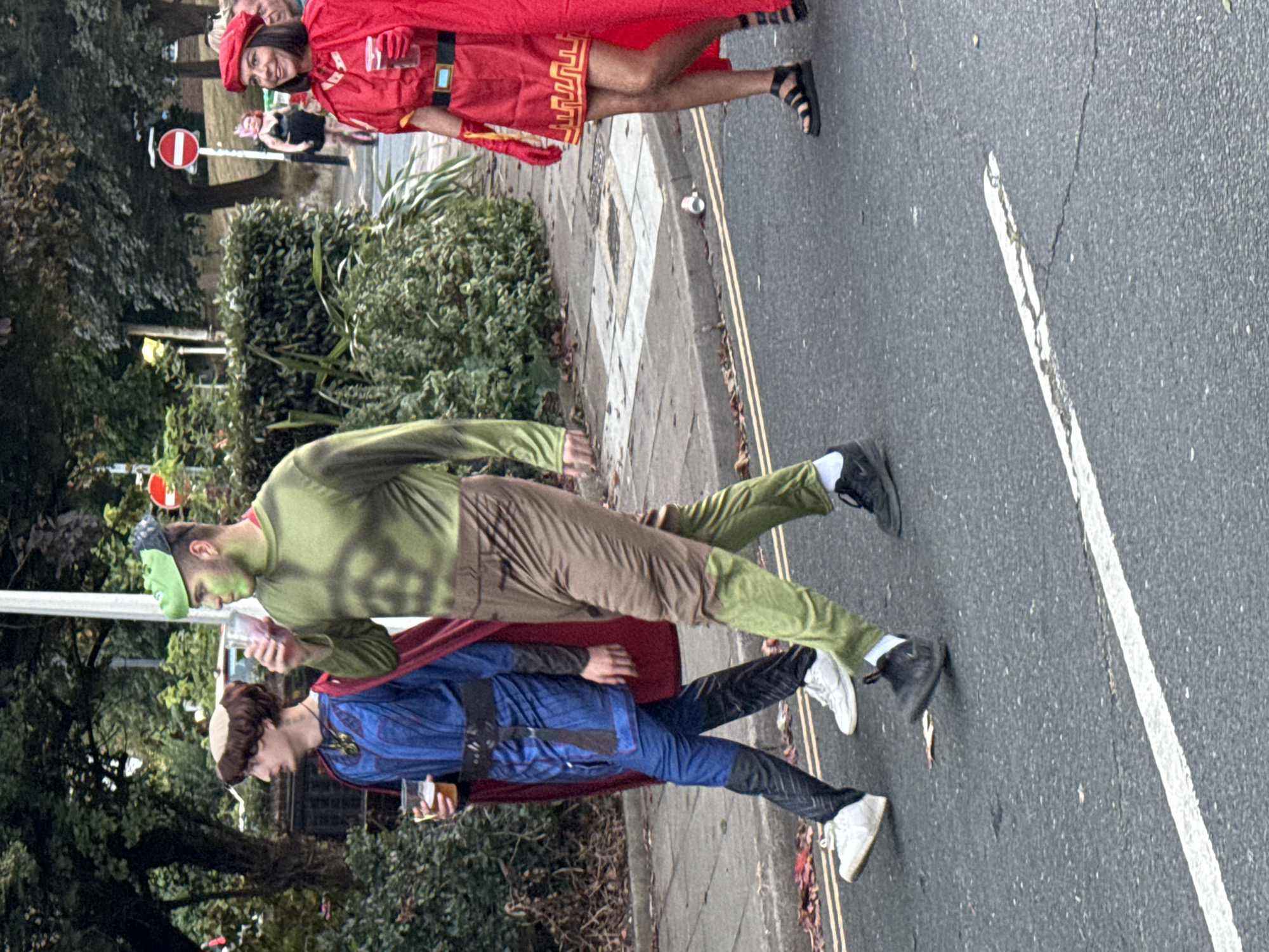 Three people are walking on a street, likely participating in a parade or event. The person in the foreground on the right is wearing a red cape, matching dress, and red gloves, with a red beret. They are holding a drink. To their left, a person in a green Hulk-like costume, complete with green face paint, is also holding a drink. The third person, on the far left, is dressed in a blue and black costume with a red cape and a brown cap, and is holding a drink. They are all walking on an asphalt road with a white line marking the lane. The background shows a sidewalk, green bushes, trees, and a glimpse of buildings and a 'no entry' sign. The scene suggests a daytime event, possibly a street festival or Halloween celebration, in St Leonards, United Kingdom.