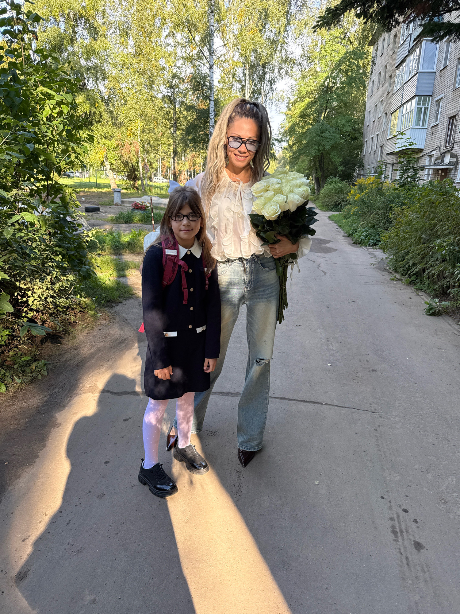 The image captures two females standing on a paved pathway in front of a multi-story brick apartment building. The female in the foreground, a young girl, wears a dark dress with white collar and cuffs, white polka-dot tights, and black shoes. She has a red backpack with straps visible over her shoulders and is holding a flower bouquet with white roses. The second female, an adult, stands behind the girl, holding the same bouquet. She is wearing a white ruffled blouse, light blue ripped jeans, and dark shoes. Both are smiling, suggesting a pleasant interaction. The environment includes trees with green and yellowing leaves, indicating an autumn setting, and a portion of the apartment building with balconies. The pathway is lined with bushes and foliage. The sun casts strong shadows on the ground, indicating it is daytime. The image appears to have been taken outdoors, in a residential area.