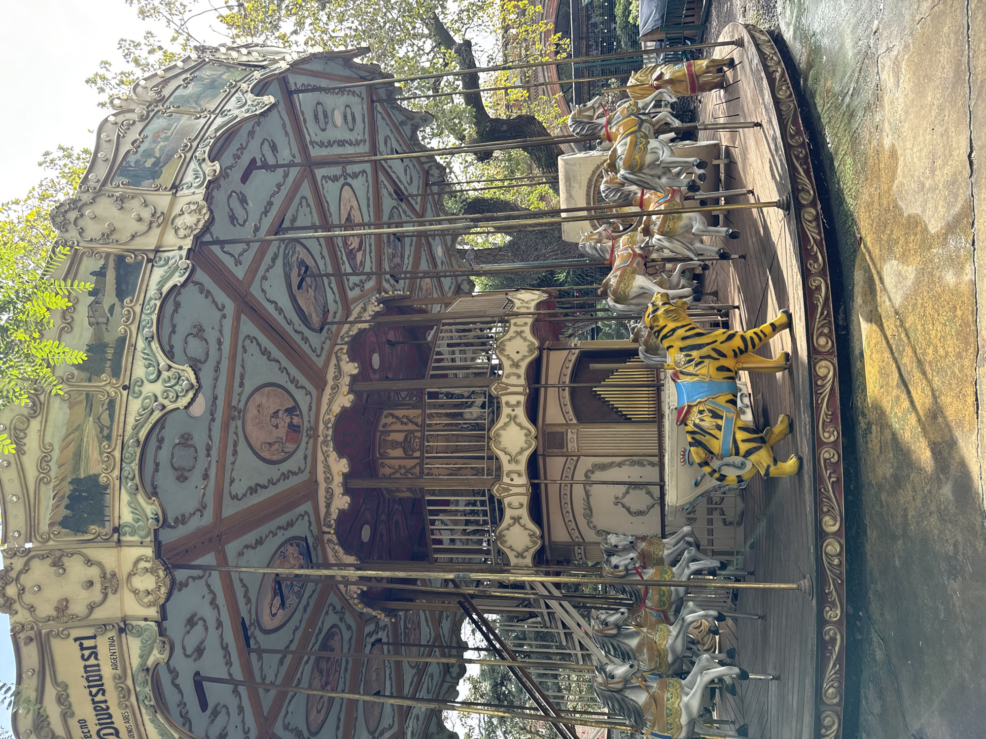 The image shows a carousel in Buenos Aires, Argentina. The carousel is ornately decorated with paintings and carvings. There are several horses and a tiger for riders. The base of the carousel is wooden with an ornamental border. The carousel is located outdoors next to a concrete area. The weather appears to be overcast. "Tecno Diversion SRL" is labeled on the top of the carousel.