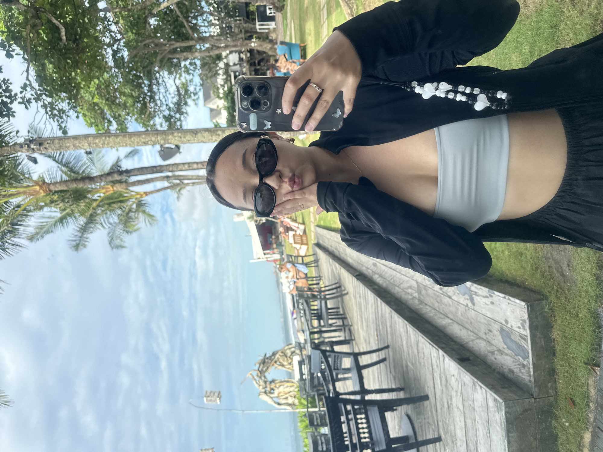 A young woman in sunglasses is taking a selfie with a smartphone on a boardwalk in North Kuta, Indonesia. She is wearing a light gray top under a black jacket, with a black beaded lanyard attached to her phone case. Her lips are pursed in a kiss and her hands are held to her cheeks. The boardwalk is lined with black chairs and tables, with a beach and ocean visible in the background. Palm trees and other greenery are visible in the background, along with structures. The sky is blue with scattered clouds.