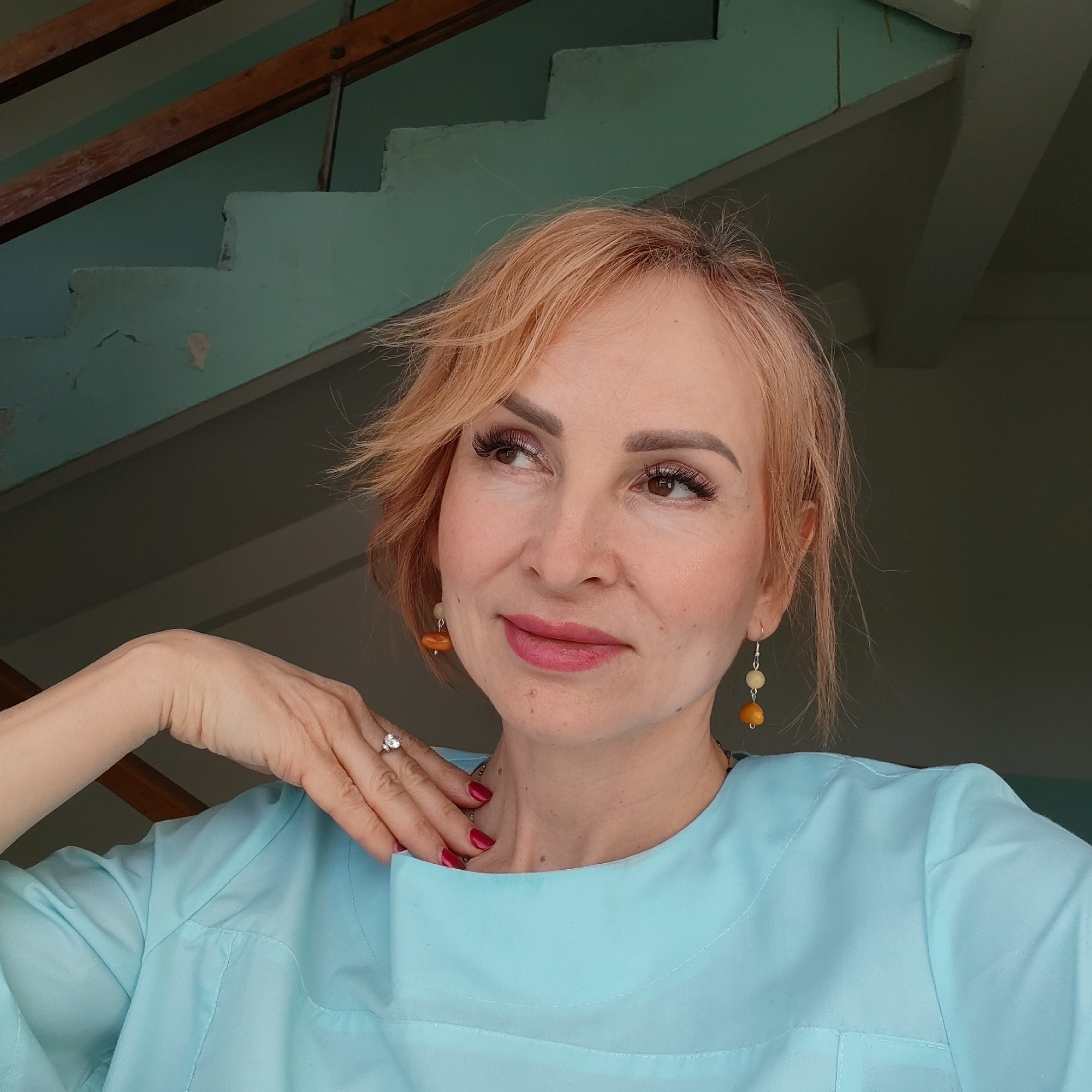 A medium shot shows a middle-aged woman with light red hair and wearing a light blue garment. She has on makeup and is wearing dangling earrings with yellow and white beads. Her left hand is raised to her neck and displays a ring on her finger, the nails are painted red. The background includes a set of painted wooden stairs with a wooden railing, and a wall painted the same light blue color as the garment, location Unknown, Unknown.