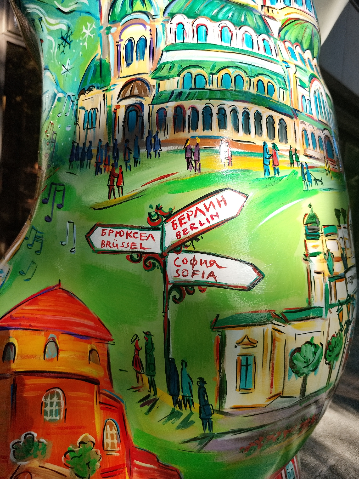 The image shows a painted object, likely a sculpture or a decorative item, depicting a cityscape scene. The style of painting is colorful and impressionistic. Architectural structures with green domes and arched windows dominate the upper portion. Small figures representing people are scattered throughout the landscape. A signpost with directions to "Брюксел/Brussel", "Берлин/Berlin", and "София/Sofia" is prominently displayed. Other architectural elements, including a building with a red roof, are visible. The overall environment is bright and sunny, with some areas in shadow. The location of the city is suggested to be a blend of European cities, incorporating landmarks or features associated with Brussels, Berlin, and Sofia.