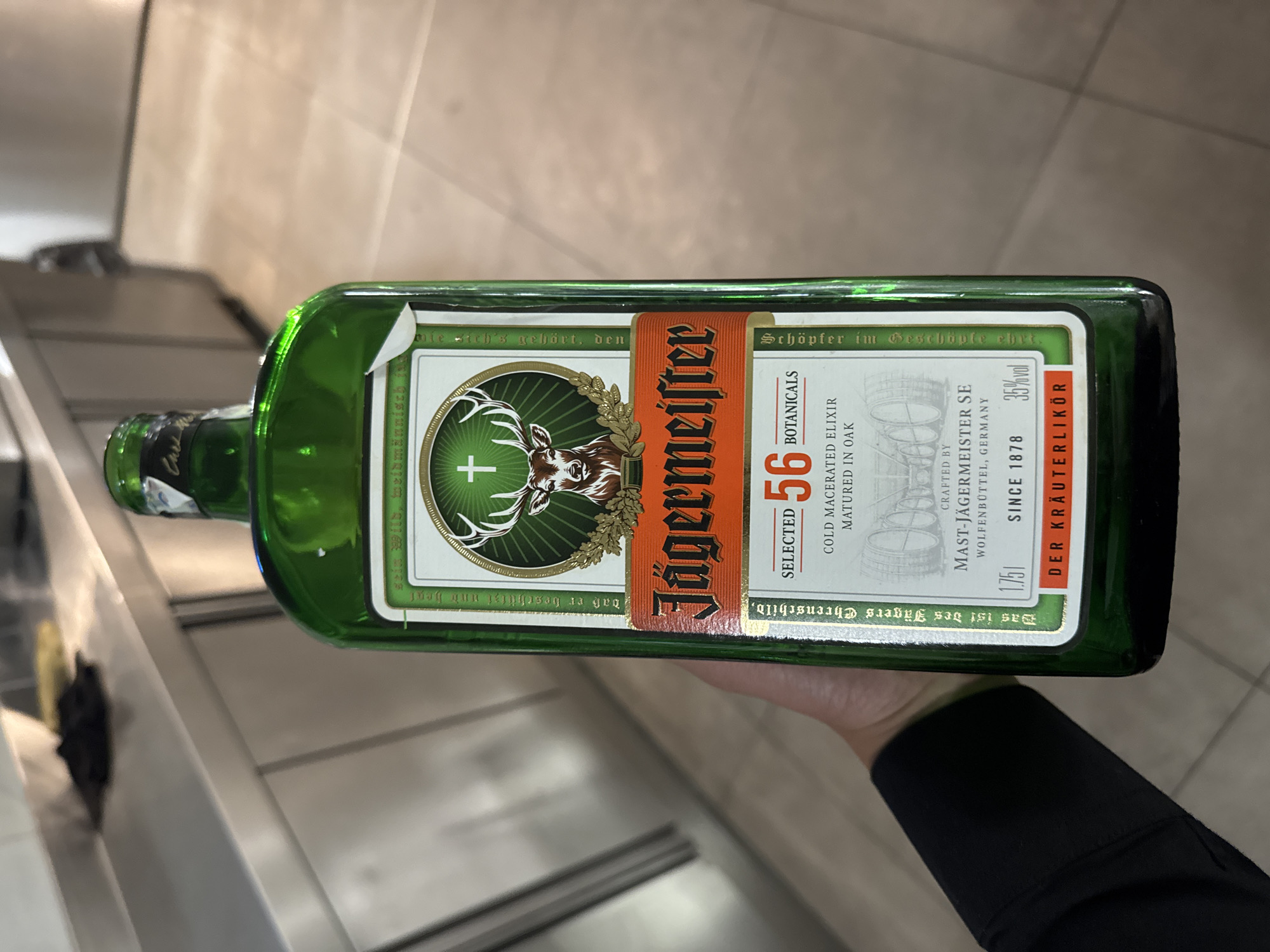 A hand holds a 1.75 liter bottle of Jägermeister. The bottle is predominantly green with an orange and white label. The label prominently features the Jägermeister logo with a deer head and cross. The label indicates it contains 56 botanicals and is a German Kräuterlikör with 35% alcohol by volume. The background is a blurred view of a metallic surface and tiled floor, suggesting an indoor location, but the specific location in Sofia, Bulgaria is impossible to ascertain from the image.