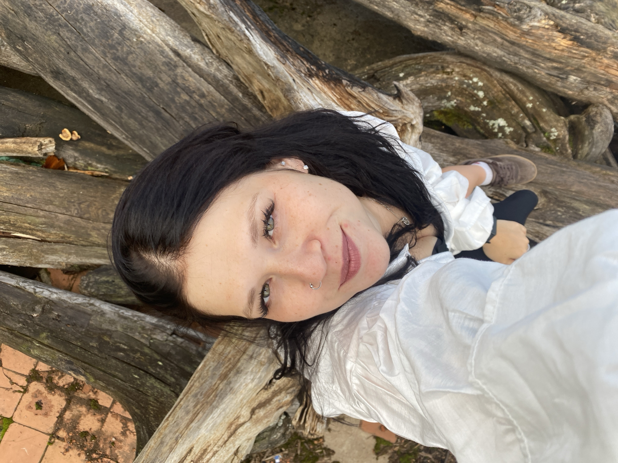 A selfie taken from above shows a young woman with long, dark hair and green eyes. She is wearing a white, loose-fitting shirt and black bottoms. The woman is positioned among large, gnarled tree trunks that appear to be part of a natural outdoor environment, possibly a park or wooded area. Some of the tree trunks are covered in moss. In the background, a section of orange tile can be seen, suggesting the presence of a paved area nearby. The lighting indicates it is daytime. There are no visible landmarks to identify the specific location within Noviy, Russia.
