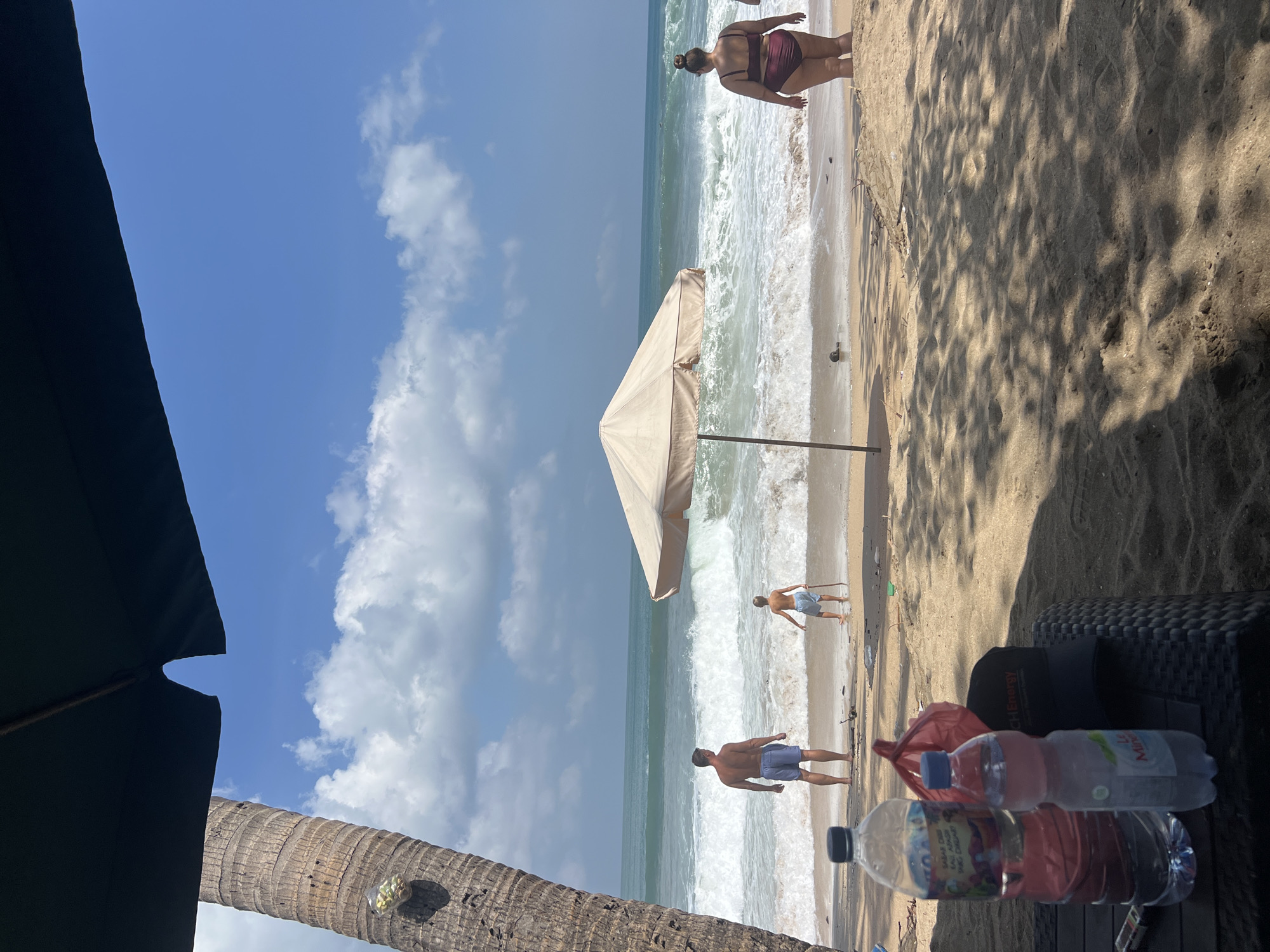 In Kuta, Indonesia, on a sandy beach, a scene unfolds under a bright, partly cloudy sky. The ocean, with its moderate waves, meets the shore, creating a lively atmosphere. A man, shirtless and wearing blue swim trunks, stands facing the water. A young boy, also in swim trunks, holds a stick and faces the ocean. To the right, a woman in a maroon two-piece swimsuit stands with her back to the camera, looking out at the sea. A beige beach umbrella provides shade over a sandy area. In the foreground, on a dark, woven table, sit two water bottles, one large clear bottle with a red bag inside and another smaller clear bottle with a pink cap. A dark baseball cap rests beside them. A portion of a palm tree trunk is visible on the left, and the shadow of an unseen canopy stretches across the sand. The overall impression is a relaxed day at the beach.