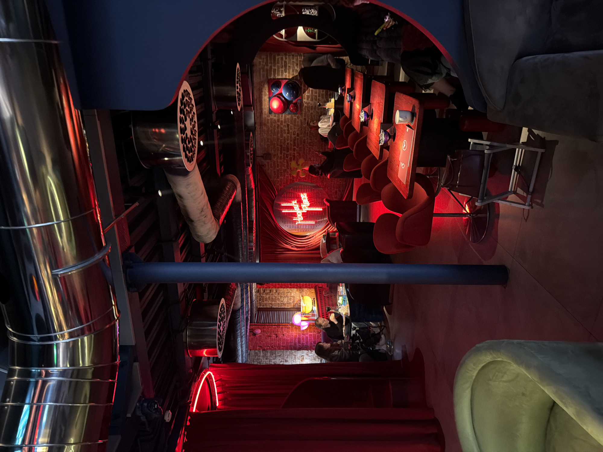 The image shows the interior of a hookah bar, likely in Moscow, Russia. The space features industrial-style decor with exposed metal pipes and circular vents, alongside brick walls and red neon lighting. Tables and chairs are arranged along one side of the room, occupied by patrons. One wall displays a neon sign reading "Hookah Place." A dark blue ceiling partially covers the view, while sofas are visible on the other side of the room.