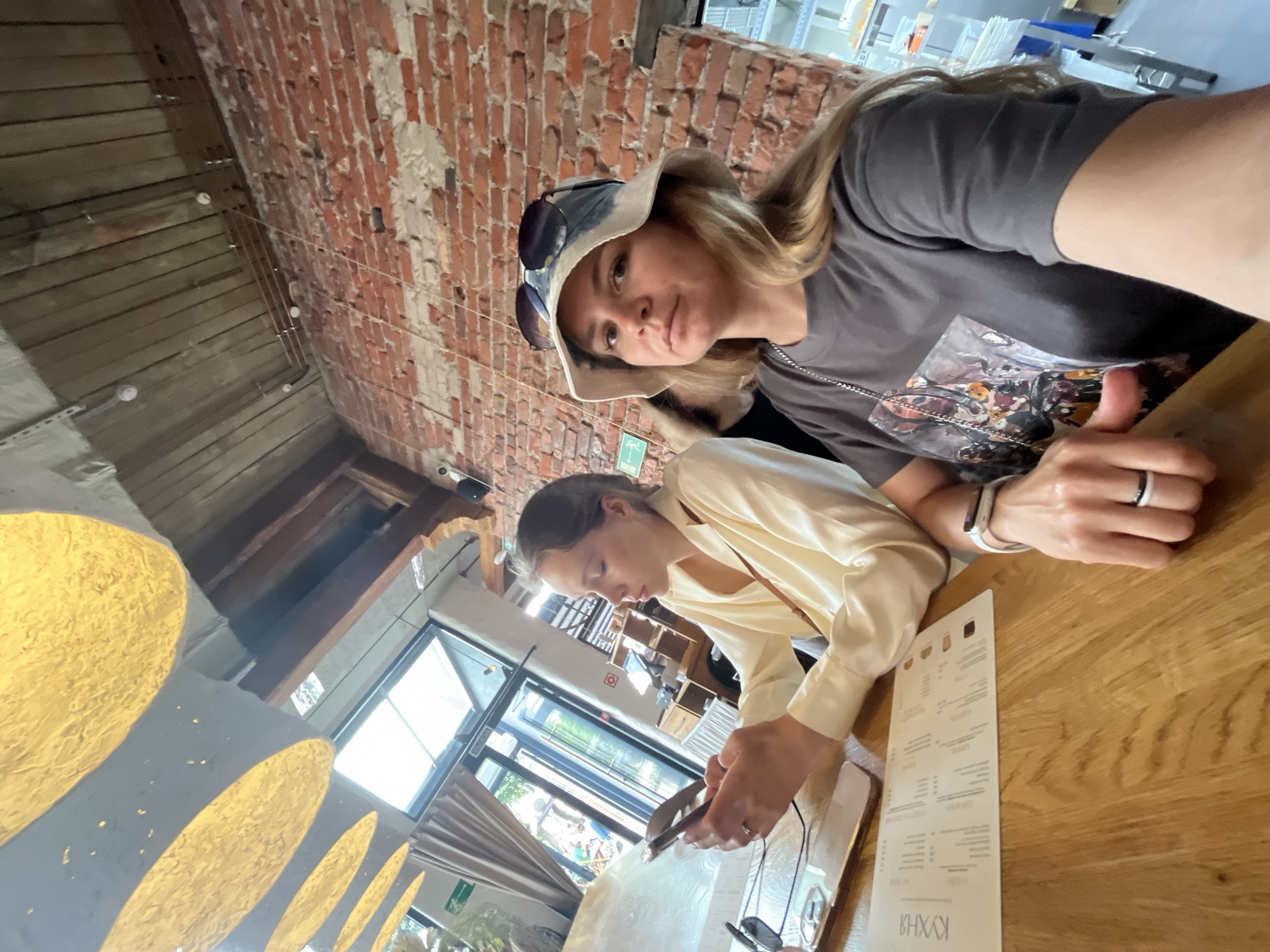 Two women are seated at a wooden table in what appears to be a restaurant with a brick wall and exposed wooden ceiling beams. The woman on the left, wearing a light-colored long-sleeved shirt, is looking at her smartphone. The woman on the right, wearing a grey t-shirt and a bucket hat, is looking towards the camera. A menu is visible on the table in front of them. The lighting suggests an indoor setting, possibly a cafe or restaurant, with large windows in the background. The overall ambiance is casual and relaxed. There is no discernible information in the image to confirm the location in Zelenogradsk, Russia.
