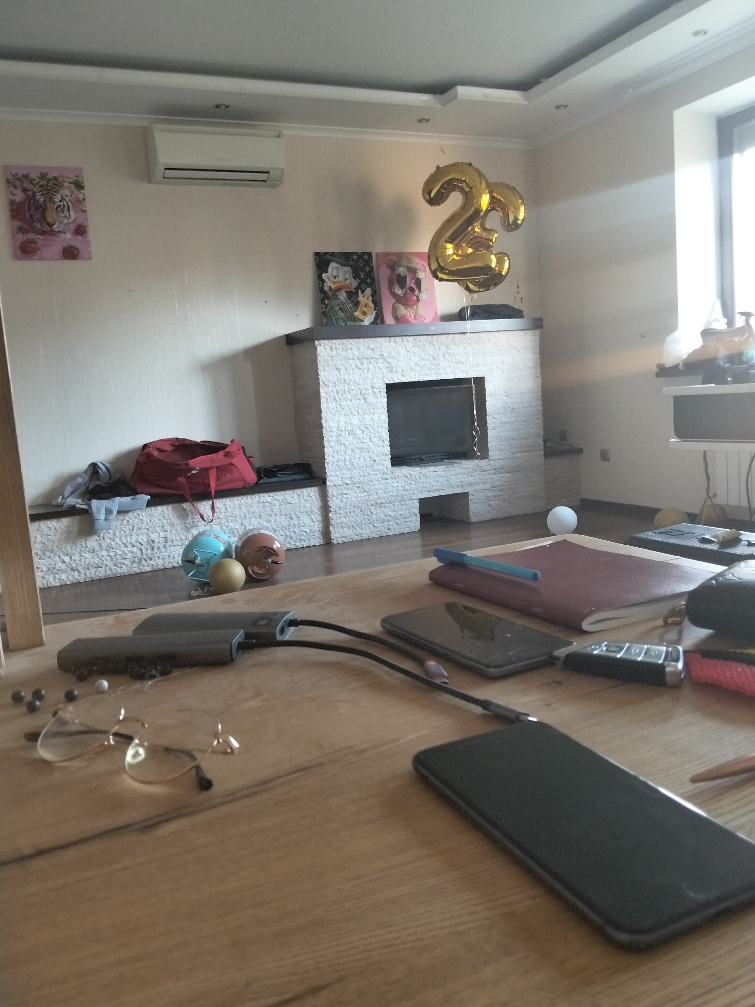 The image depicts an indoor scene, likely a living room or den, with a focus on a wooden table in the foreground. On the table, several electronic devices are visible, including two dark-colored smartphones, one of which is plugged into a charger. A set of keys and a pair of eyeglasses with gold frames are also present on the table. In the background, a fireplace with a stone facade is visible, adorned with framed artwork and large gold balloons shaped like the number '2'. To the left of the fireplace, there's a shelf holding a red duffel bag and some clothing. An air conditioning unit is mounted on the wall above the shelf. The room is lit by natural light streaming in from a large window on the right side of the image.
