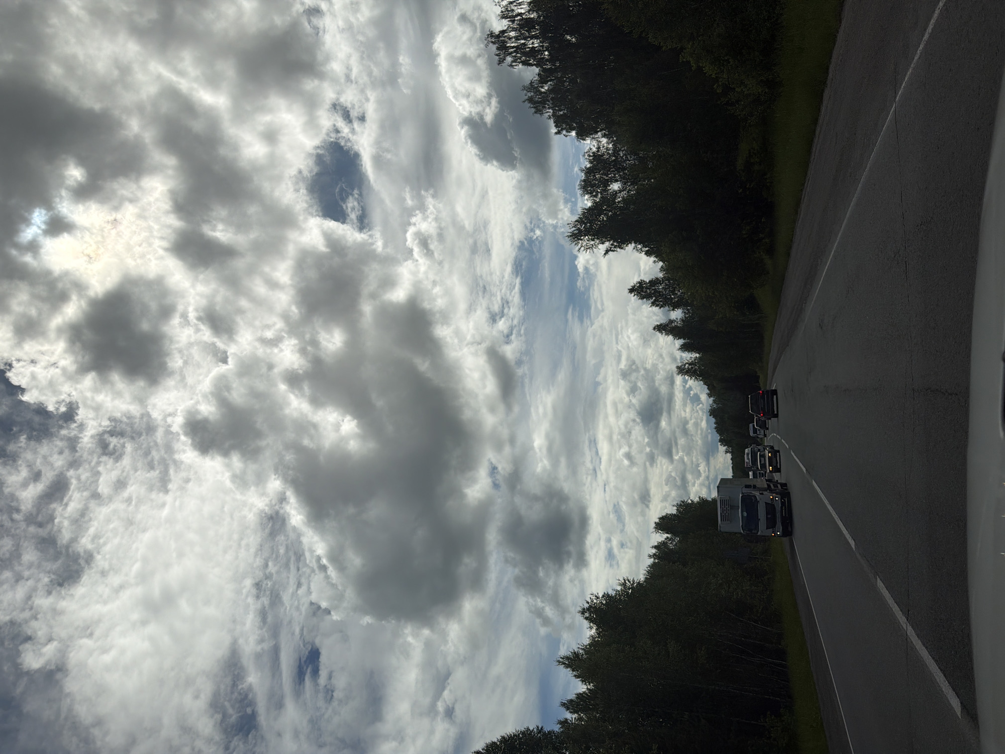A grey, overcast sky with scattered white clouds dominates the upper portion of the image. The sun is partially visible through a gap in the clouds, casting a bright glow. Below the sky, a dense, dark green forest lines both sides of a two-lane asphalt road. The road stretches into the distance, with several vehicles traveling in the same direction. In the foreground, a white truck is visible, followed by several cars. The scene appears to be a highway or a major road outside of a city or town. The location is identified as Poletaevo 1th, Russia.