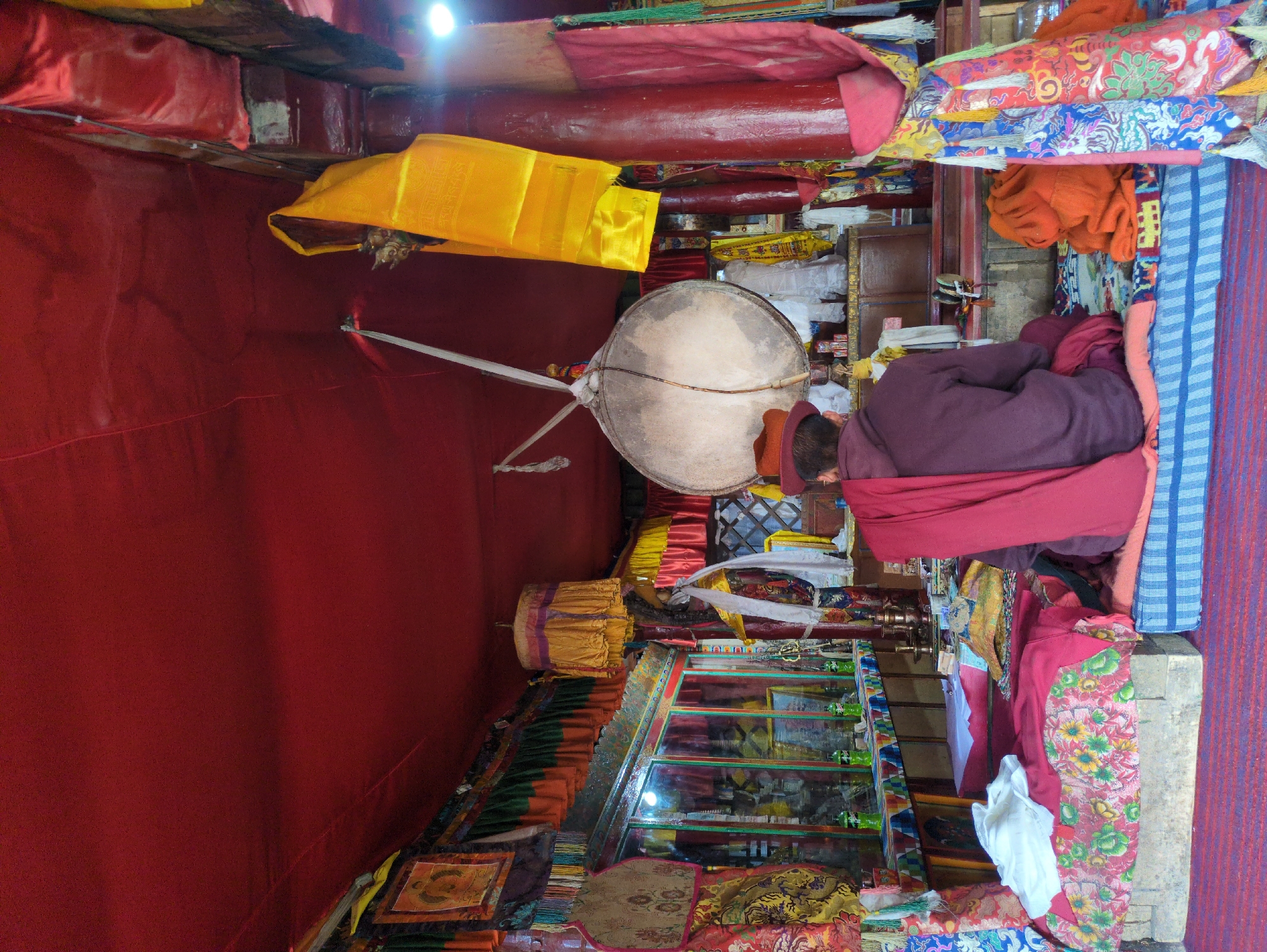 The image depicts the interior of a dimly lit room, possibly a temple or monastery, in Yulang, India. The room is adorned with red fabric draped from the ceiling and walls. In the foreground, a person wearing a maroon robe and a red hat is seated facing away from the camera, meditating or engaged in prayer. They are sitting on a colorful, patterned cushion placed on a raised platform.
The room contains various religious objects and decorations. A large, circular drum hangs from a support structure in the center of the room. Colorful fabrics, including yellow and red streamers, are draped around the space. On the left side of the room, there is a display case or shelf filled with bottles and other items. The overall ambiance suggests a place of worship or spiritual practice, with a rich cultural and religious aesthetic.