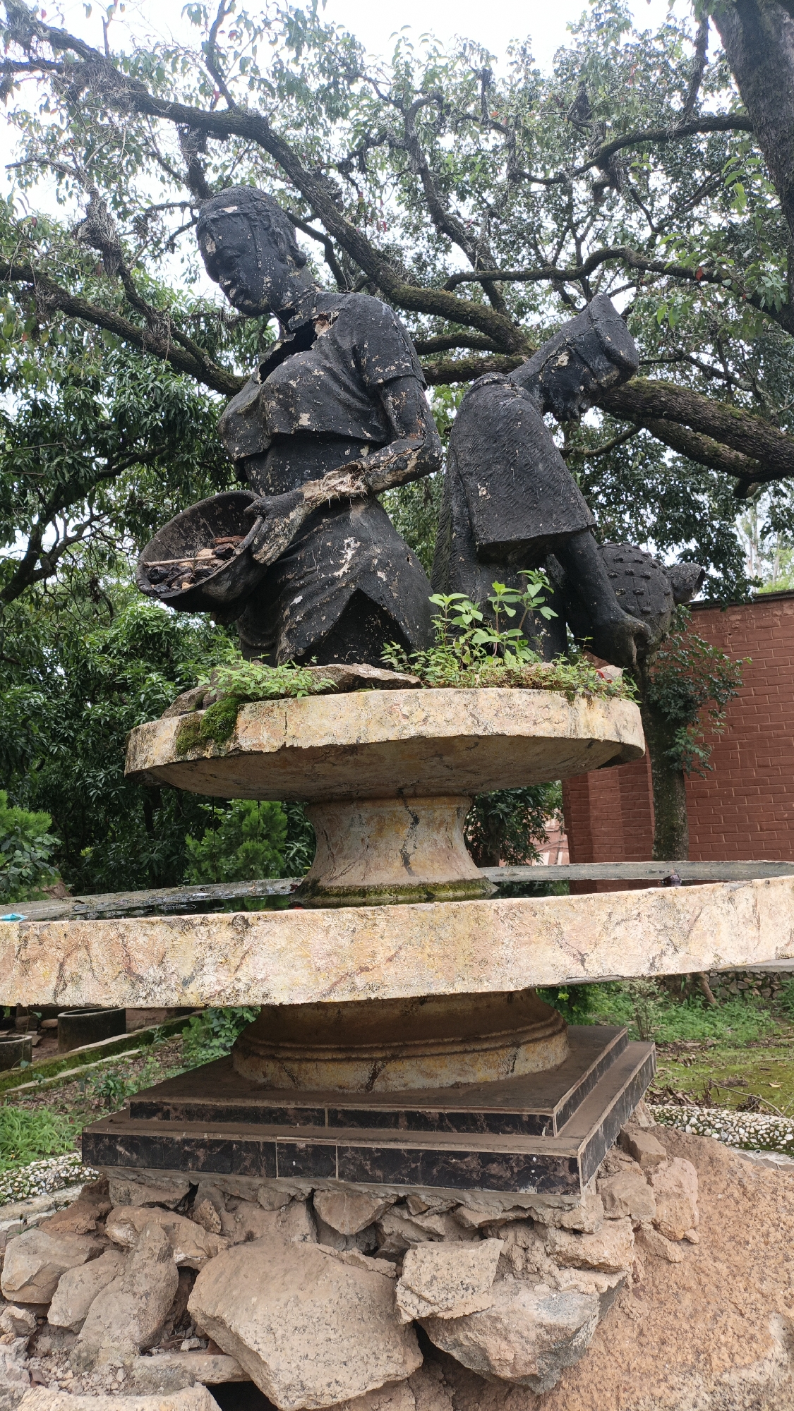 This is a medium shot, outdoor, ground-level photograph of a weathered, dark-colored sculpture depicting two figures in traditional clothing, standing on a multi-tiered, stone-like pedestal. The sculpture and pedestal are centrally located in the frame, with lush green foliage and gnarled tree branches overhead, suggesting a park or garden setting. In the background, a red brick wall is visible, with some greenery and other structures peeking through the trees. The sculpture itself appears to be made of a rough material, possibly concrete or stone, and shows signs of age and wear, with some areas appearing chipped or incomplete. The two figures are positioned side-by-side, with their heads bowed and looking downwards. The figure on the left is holding a shallow basket filled with what appears to be dried leaves or debris. The figure on the right is also holding an object, partially obscured by their clothing. Small green plants are growing around the base of the sculpture on the pedestal, adding a touch of life to the scene. The base of the pedestal is a dark, tiered structure, surrounded by a pile of rough, earthy-toned rocks. The overall impression is one of a historical or artistic monument situated in a natural environment. This sculpture is located at the National Museum Jos, Plateau State, Nigeria, a significant cultural heritage site known for its diverse collections and outdoor displays, including historical sculptures and artifacts.