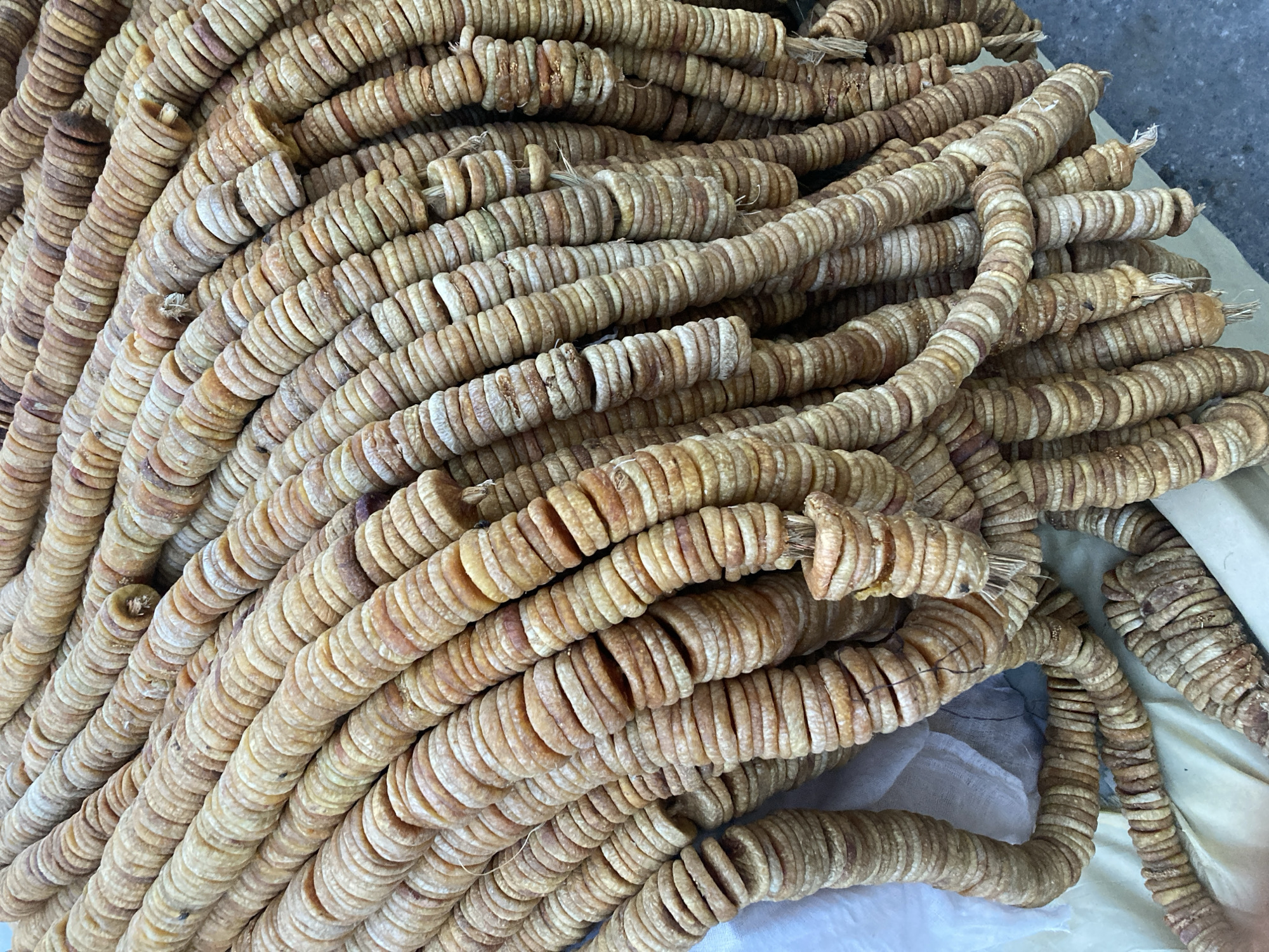 A close-up photograph depicts a large pile of strings of dried figs. The figs are circular slices threaded together. The colors range from pale beige to brown. The background is slightly blurred, showing a white cloth. Based on the query, the image is likely taken in Quetta City Tehsil, Pakistan.