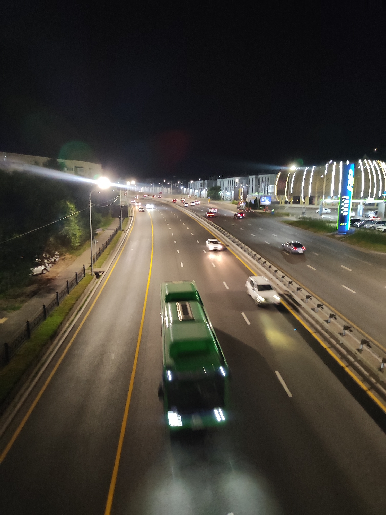 The image captures a nighttime view of a multi-lane highway in Almaty, Kazakhstan. The highway is illuminated by streetlights and the headlights and taillights of vehicles. In the foreground, a green bus is in motion, its headlights casting a bright beam. Several cars are visible on the highway, some in motion and some parked on the side. On the left side of the highway, there is a pedestrian walkway with a fence and trees. On the right side, a brightly lit commercial area with a sign that appears to read "RP" is visible, suggesting it might be a gas station or a business complex. The sky is dark, indicating it is late at night. The overall scene depicts a typical urban highway environment during nighttime.