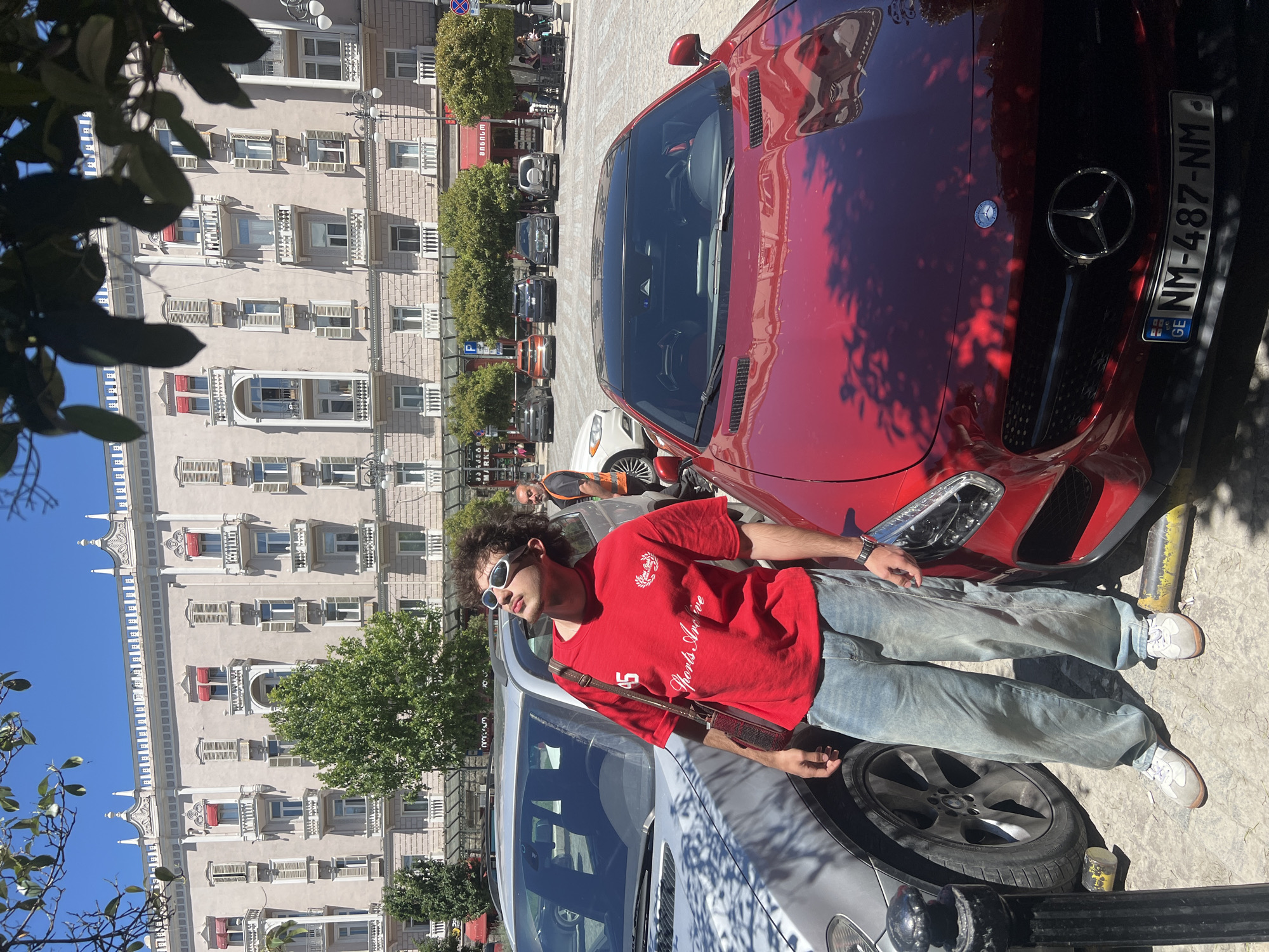 A young man with curly hair is standing between two parked cars, a silver SUV and a red sports car, in an outdoor urban setting. He is wearing a red t-shirt with white lettering and graphics, light blue baggy jeans, and white sneakers. He is also wearing white sunglasses and has a brown bag slung over his shoulder. The man is looking upwards with his mouth slightly open. In the background, a large, multi-story building with classical architectural details stands behind a row of trees. Several other cars are parked in front of the building. The ground is paved with cobblestones. The scene appears to be in a public square in Tbilisi, Georgia, as indicated by the license plate on the red sports car which reads "GE NM-487-NM". The sky is clear and blue, and the sun is casting shadows.