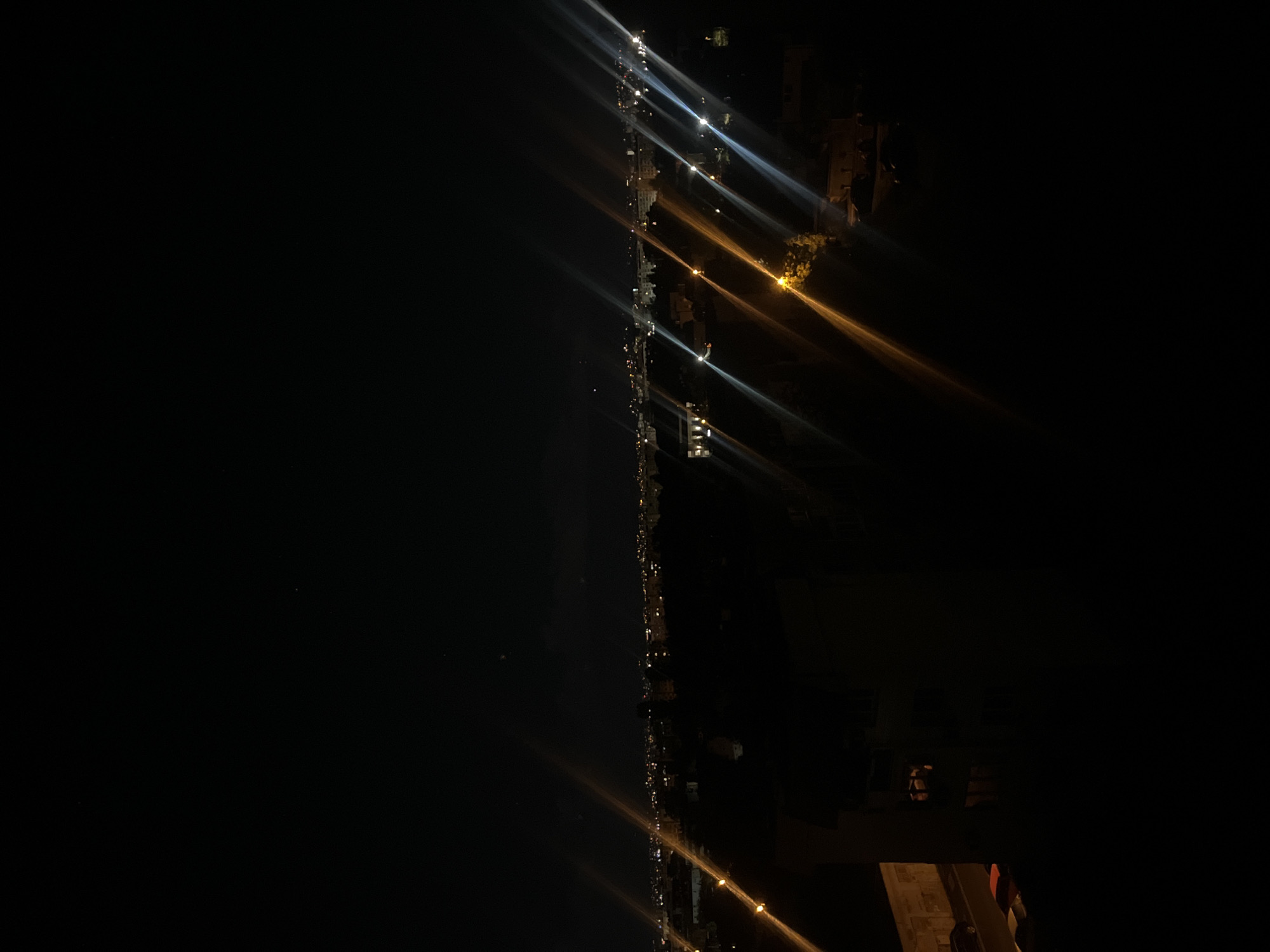 The image appears to be a nighttime view from a high vantage point, likely a building, in what is suggested to be Sofia, Bulgaria. A row of lights, possibly streetlights, extends into the distance, with flares of light emanating from them. There is a dark, undefined area on the left side of the image. On the right side, there are blurry, darker shapes that may represent buildings or other structures. The environment seems urban.