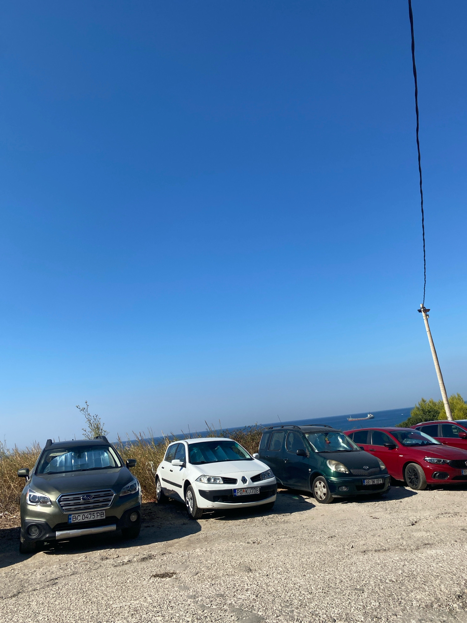 The image depicts a row of parked cars on a gravel surface near a body of water, likely the Adriatic Sea. The sky is a clear, vibrant blue. In the foreground, a dark green Subaru Outback is parked. To its right, a white Renault Megane is parked, followed by a dark green Toyota Yaris Verso, a red Fiat Tipo, and a red vehicle whose make and model are less clear. A distant ship is visible on the water. A utility pole with a wire extending upwards is present on the right side of the image. The scene appears to be a parking area with a view of the sea, possibly on a coastal road or overlook. The location is identified as Bar, Montenegro.