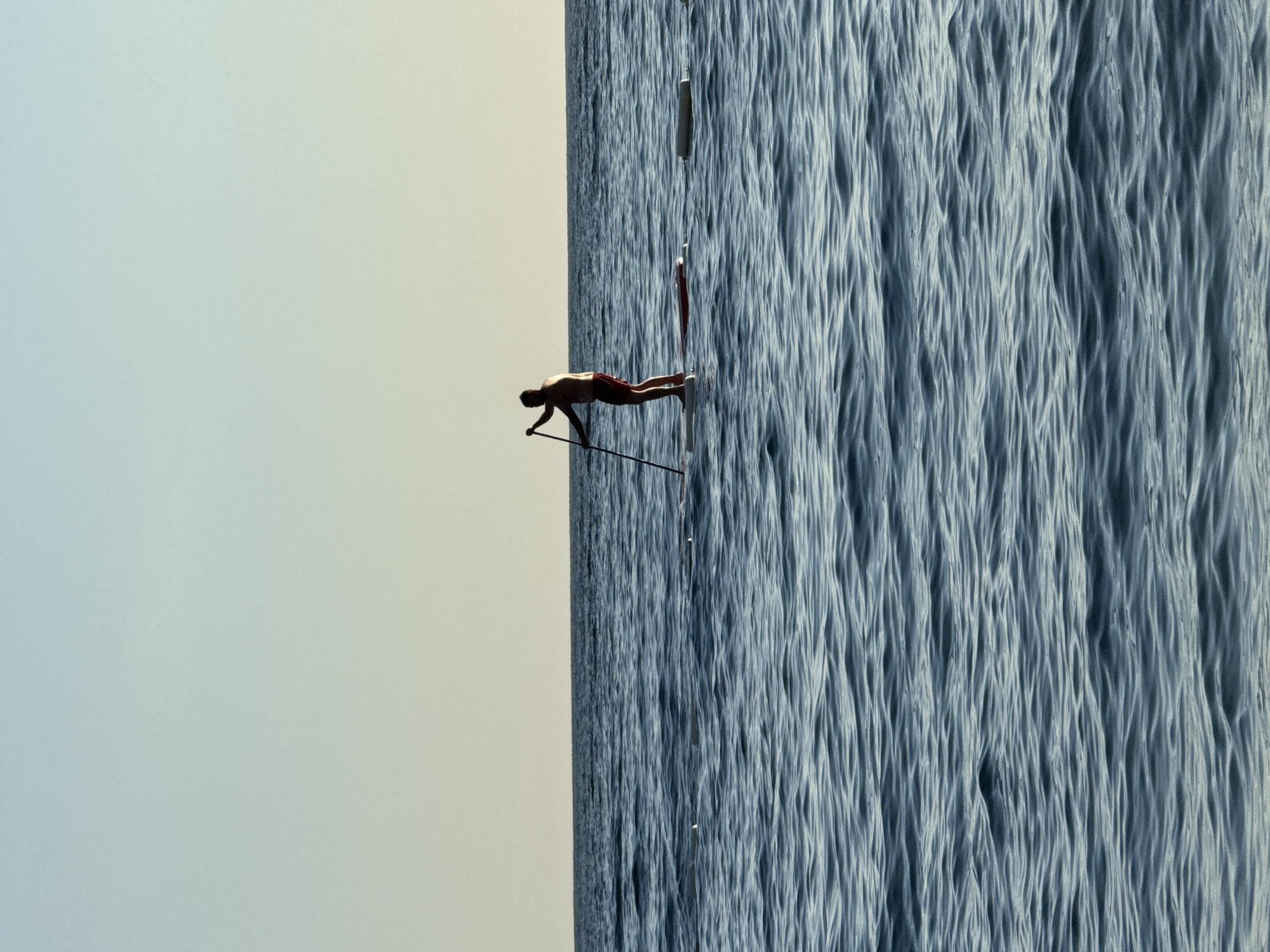 A solitary person is paddleboarding on a calm sea under a clear, gradient sky, transitioning from pale blue to a pale yellow near the horizon. The person, silhouetted against the sky, stands on a paddleboard, holding a paddle. Two white buoys are visible in the water to the right of the paddleboarder. The scene suggests an early morning or late afternoon setting.